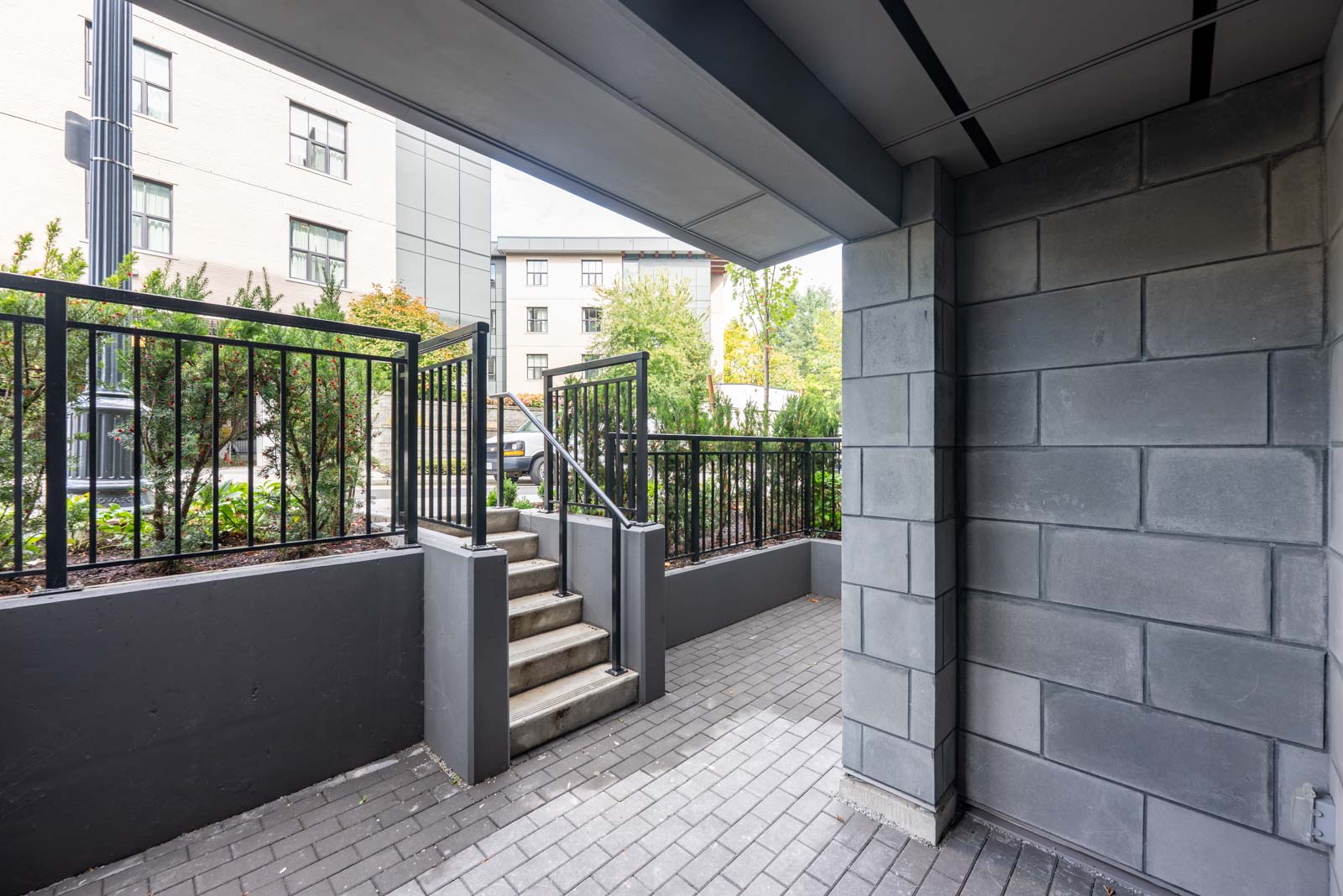 View of an outdoor stairway with metal railings, gray brick walls, and a paved walkway leading to a landscaped area and buildings in the background.