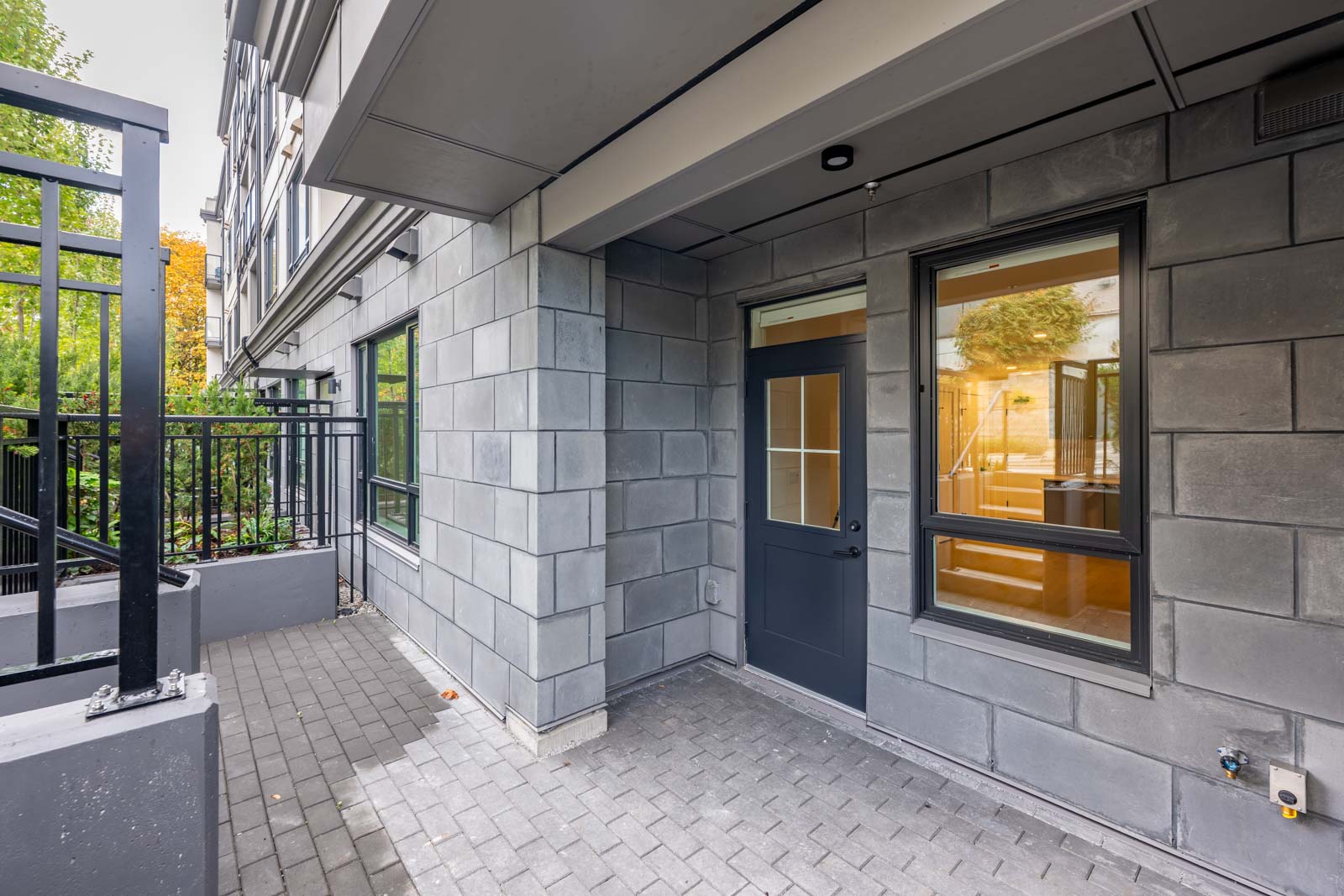Ground-level exterior view of a modern apartment patio with gray brick walls, large windows, and a black-framed glass door.
