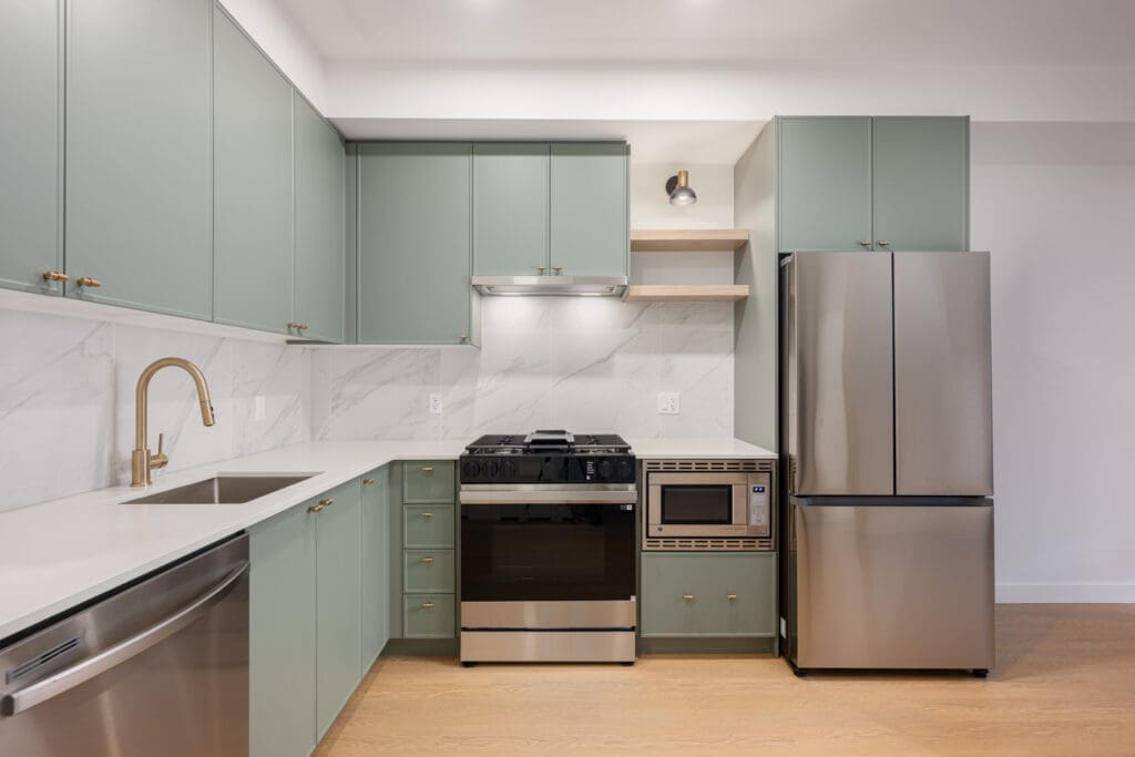 Modern kitchen with light green cabinets, stainless steel appliances, marble backsplash, and wood flooring. Features a sink, dishwasher, stove, microwave, and refrigerator.