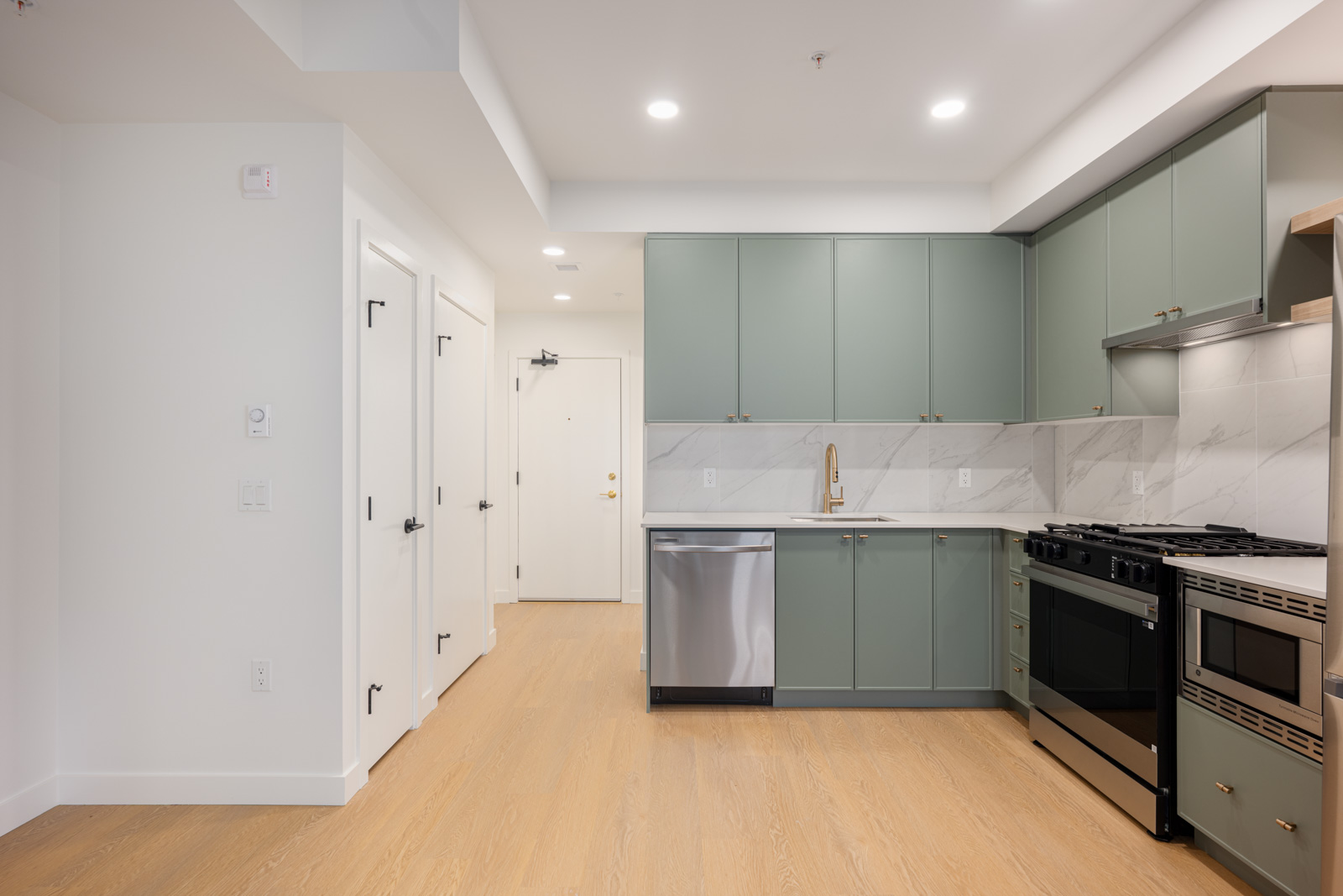 Modern kitchen with green cabinets, stainless steel appliances, light wood flooring, and white marble backsplash. Two closed doors and an entrance door are visible in the background.