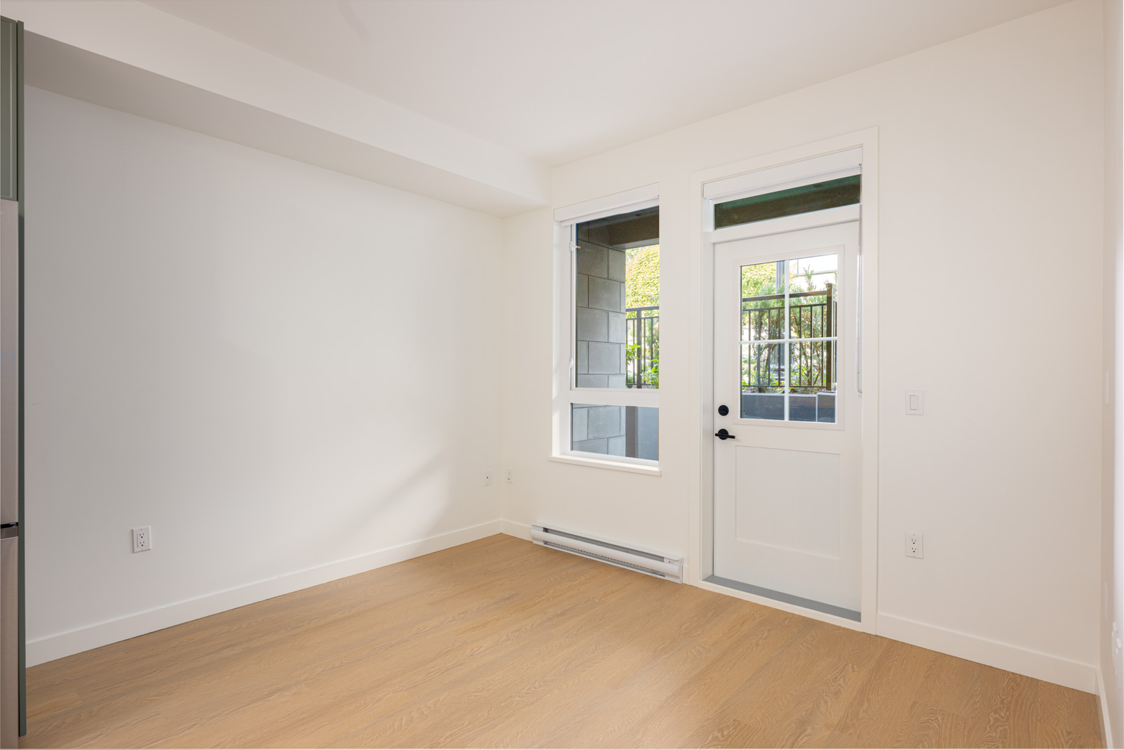 A bright, empty room with light wood flooring, white walls, a window, and a white door leading outside. Sunlight enters through the window and door.