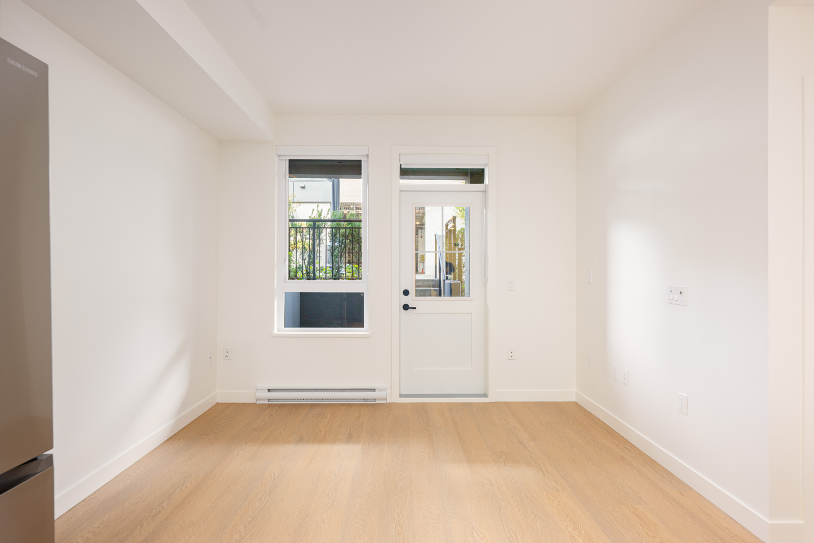 A small, empty room with light wood flooring, white walls, a window, and a door leading to an outdoor area. Part of a refrigerator is visible on the left.
