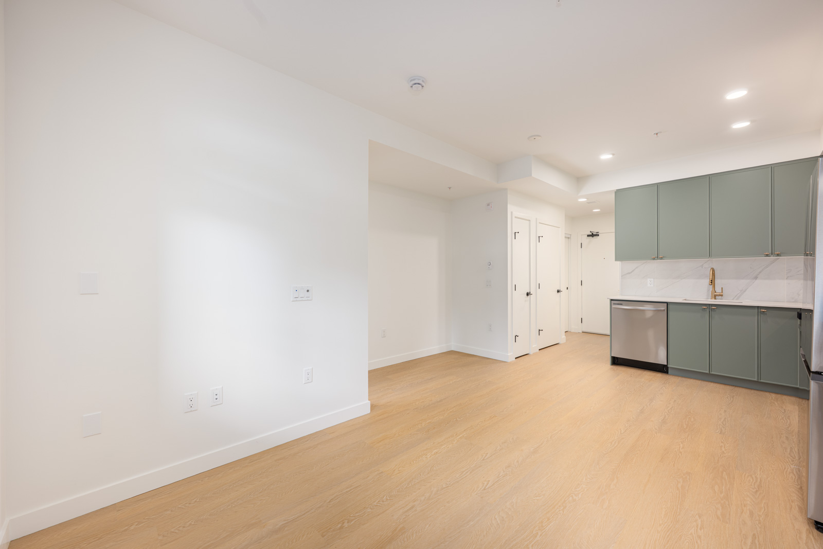 Modern, unfurnished apartment kitchen and living area with light wood floors, white walls, green cabinets, marble backsplash, and stainless steel appliances.