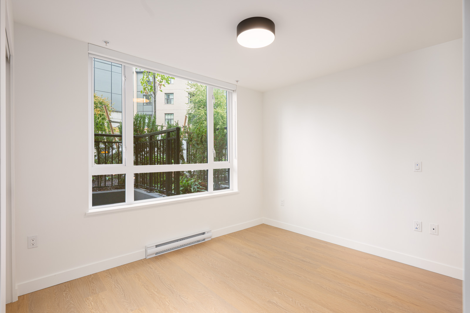 Empty, well-lit room with white walls, large window, wood flooring, ceiling light, and baseboard heater. Trees and building visible outside the window.