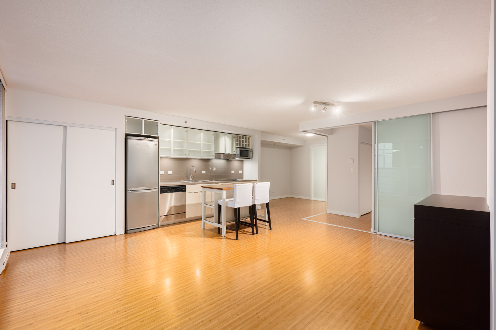 Modern open-concept apartment with light wood floors, compact kitchen, stainless steel appliances, a small dining area, and sliding glass doors.
