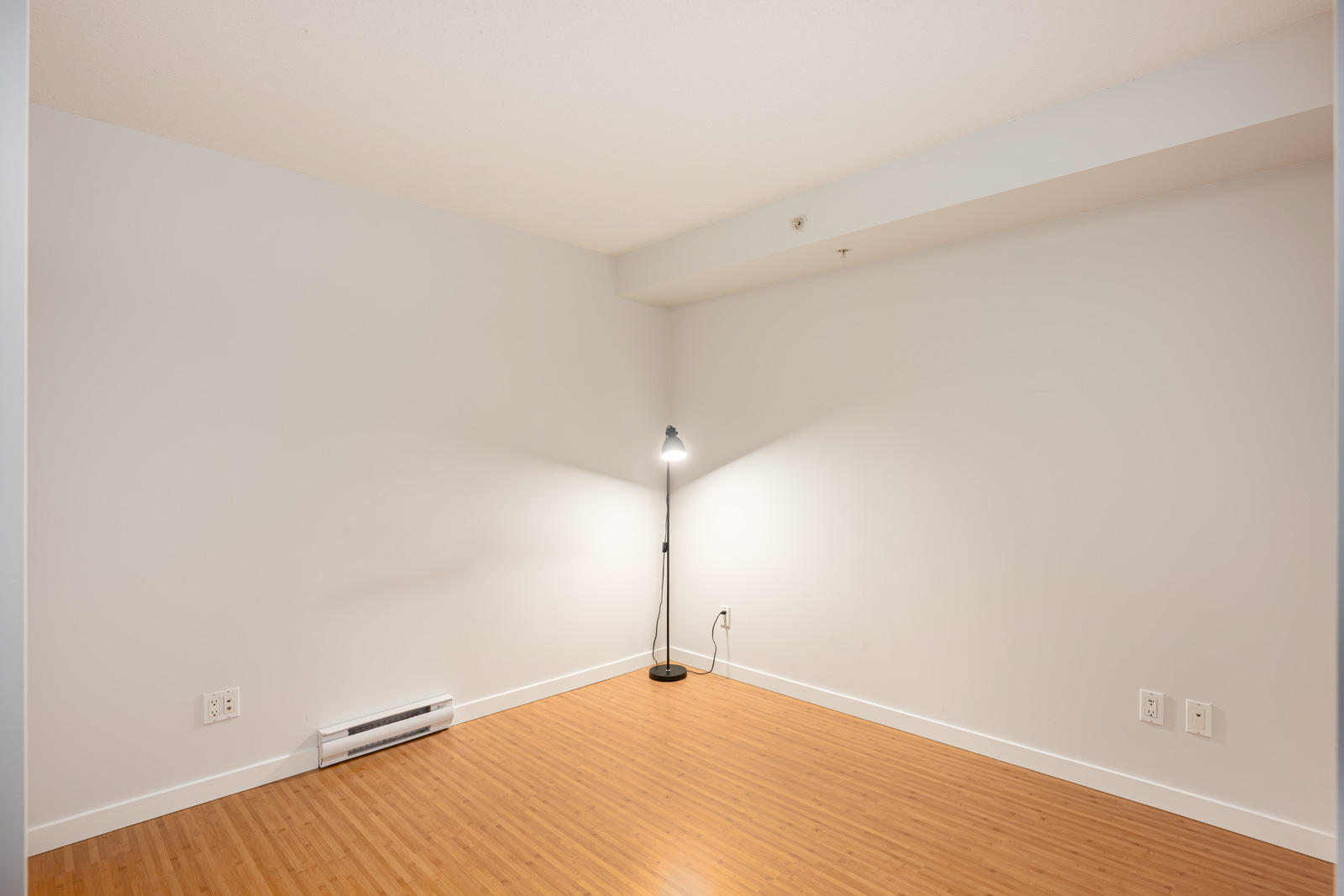 Empty room with light gray walls, light wood flooring, a single floor lamp in the corner, electrical outlets, and a baseboard heater visible.