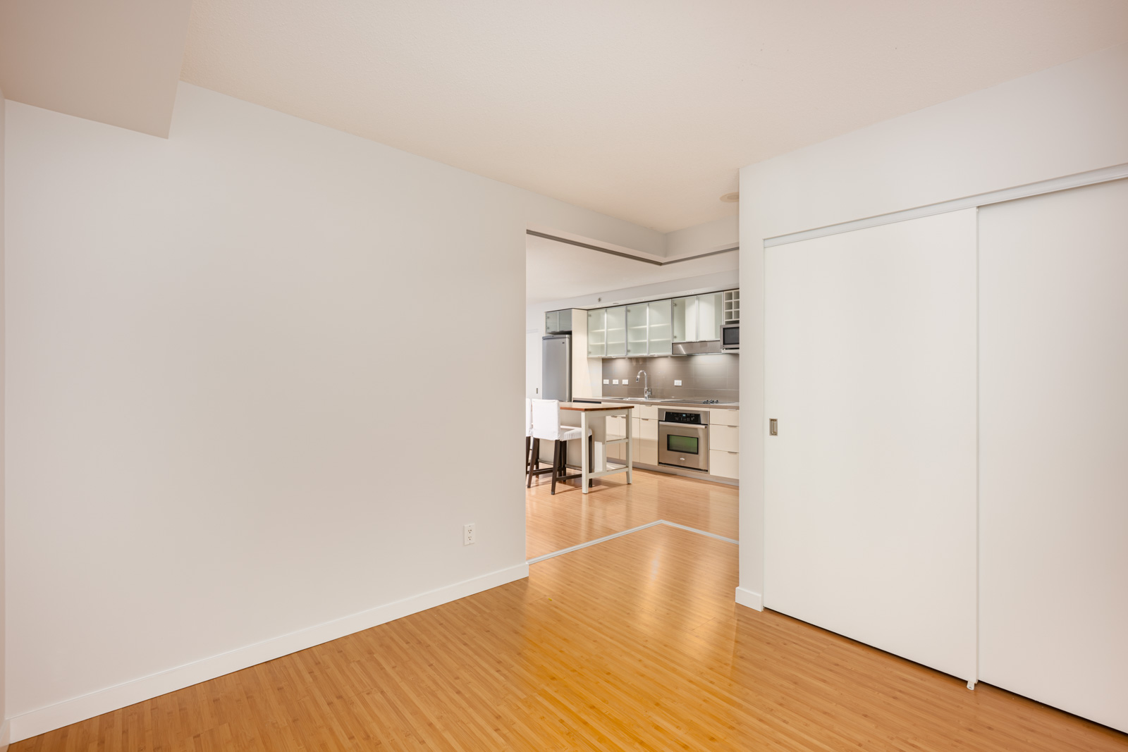 Unfurnished room with light-colored walls and wood flooring, leading to a modern kitchen with white cabinets and stainless steel appliances.