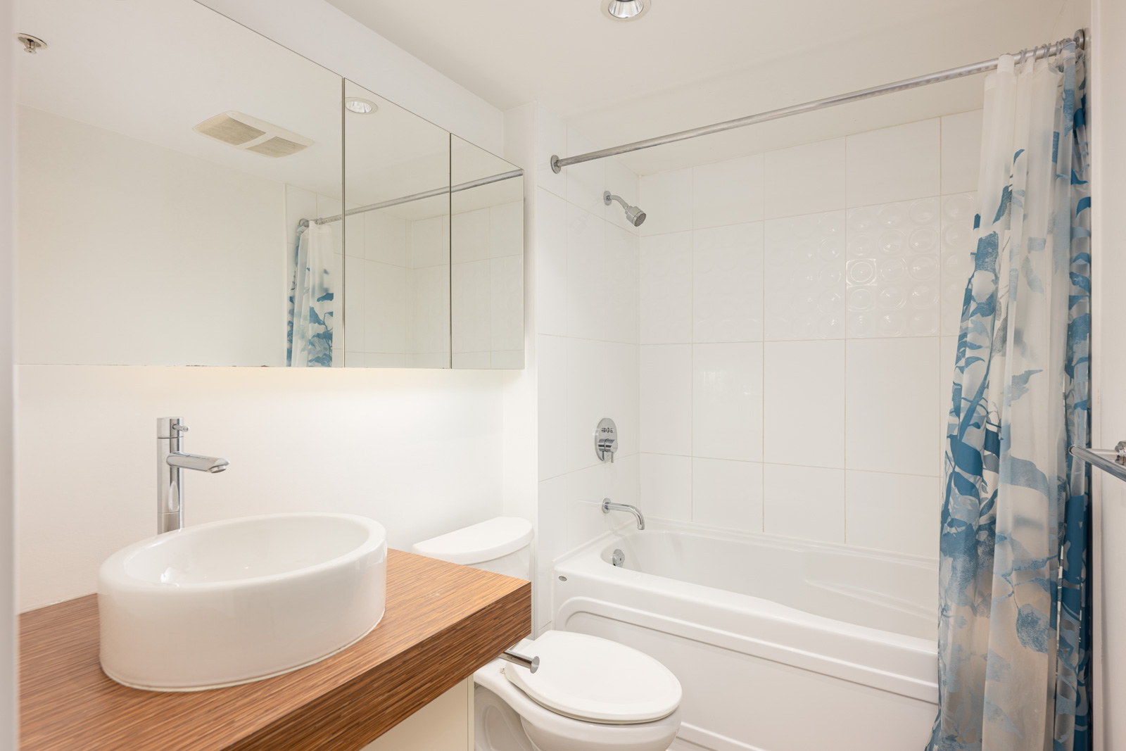 Modern bathroom with a round white sink on a wooden countertop, a toilet, a bathtub with a shower, and a floral shower curtain. Mirrored cabinets are above the sink.