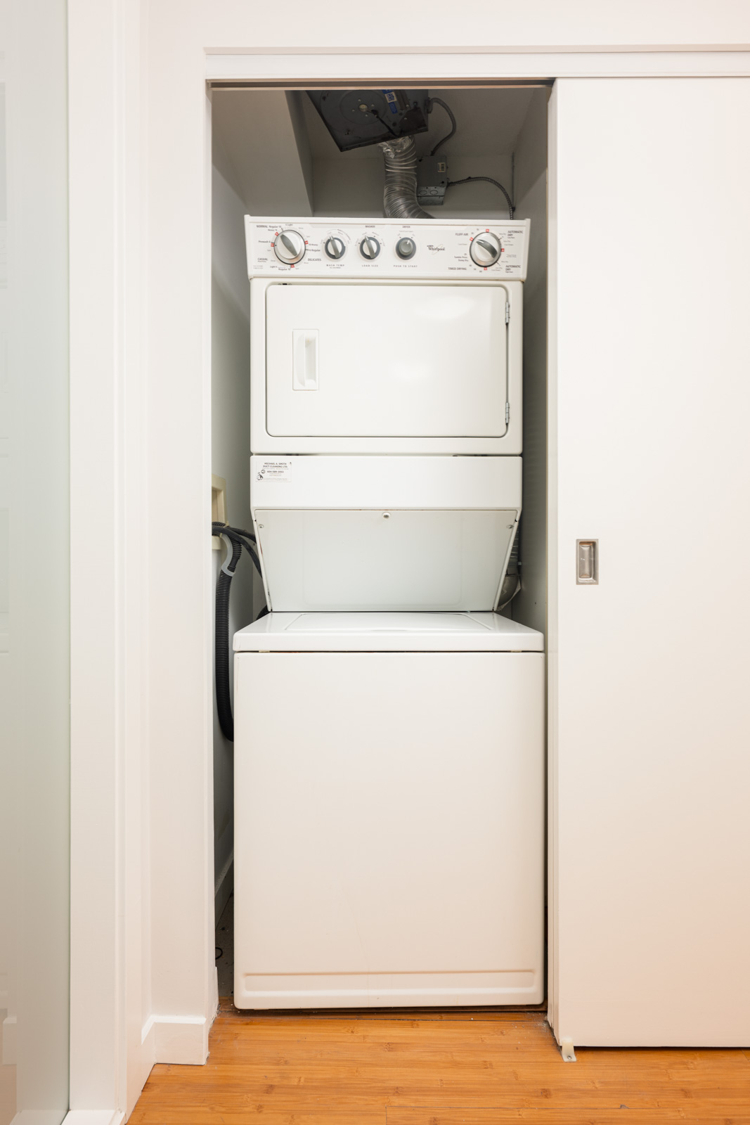 A stacked washer and dryer unit is installed in a small closet with a sliding door, next to a wooden floor.