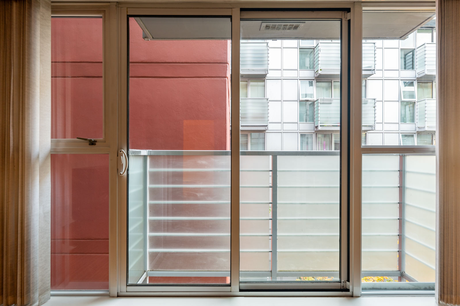 View through a glass sliding door to a small balcony with frosted glass panels; neighboring apartment buildings are visible outside.