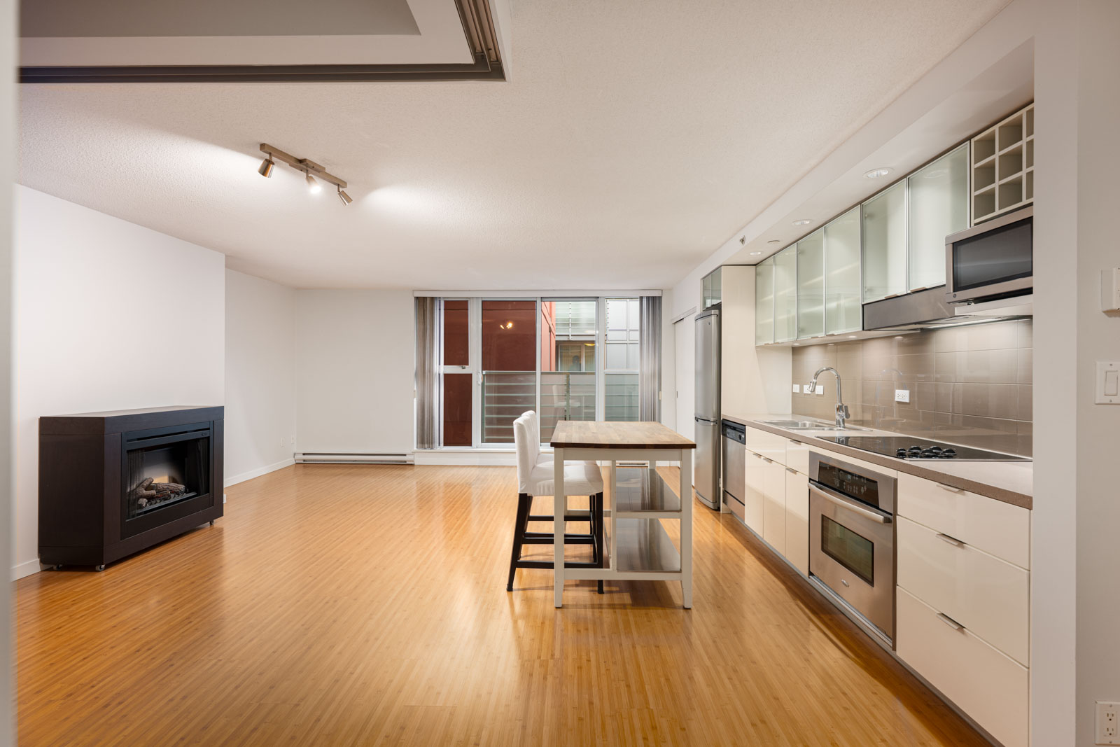 Modern open-concept kitchen and living area with wood floors, white cabinets, island with two chairs, large window, and built-in fireplace.