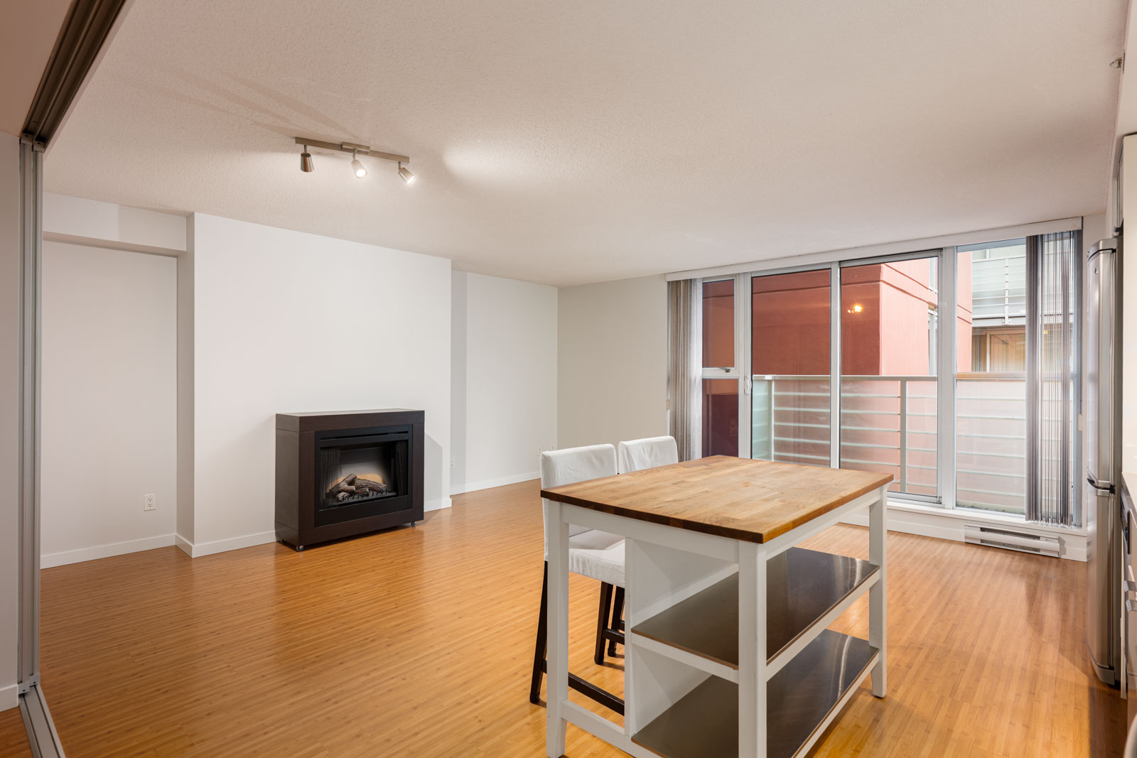 Unfurnished modern living space with hardwood floors, a fireplace, large windows, and a kitchen island with two chairs. Balcony visible through glass doors.