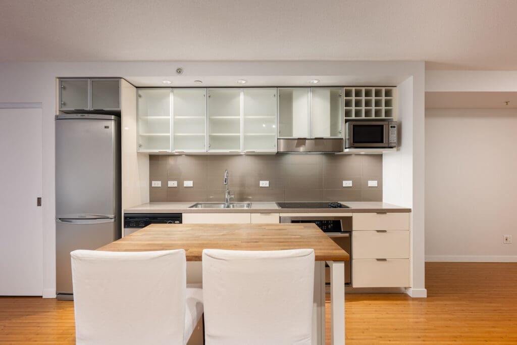 Modern kitchen with white cabinets, stainless steel fridge, microwave, open shelving, and a wooden dining table with two white chairs.