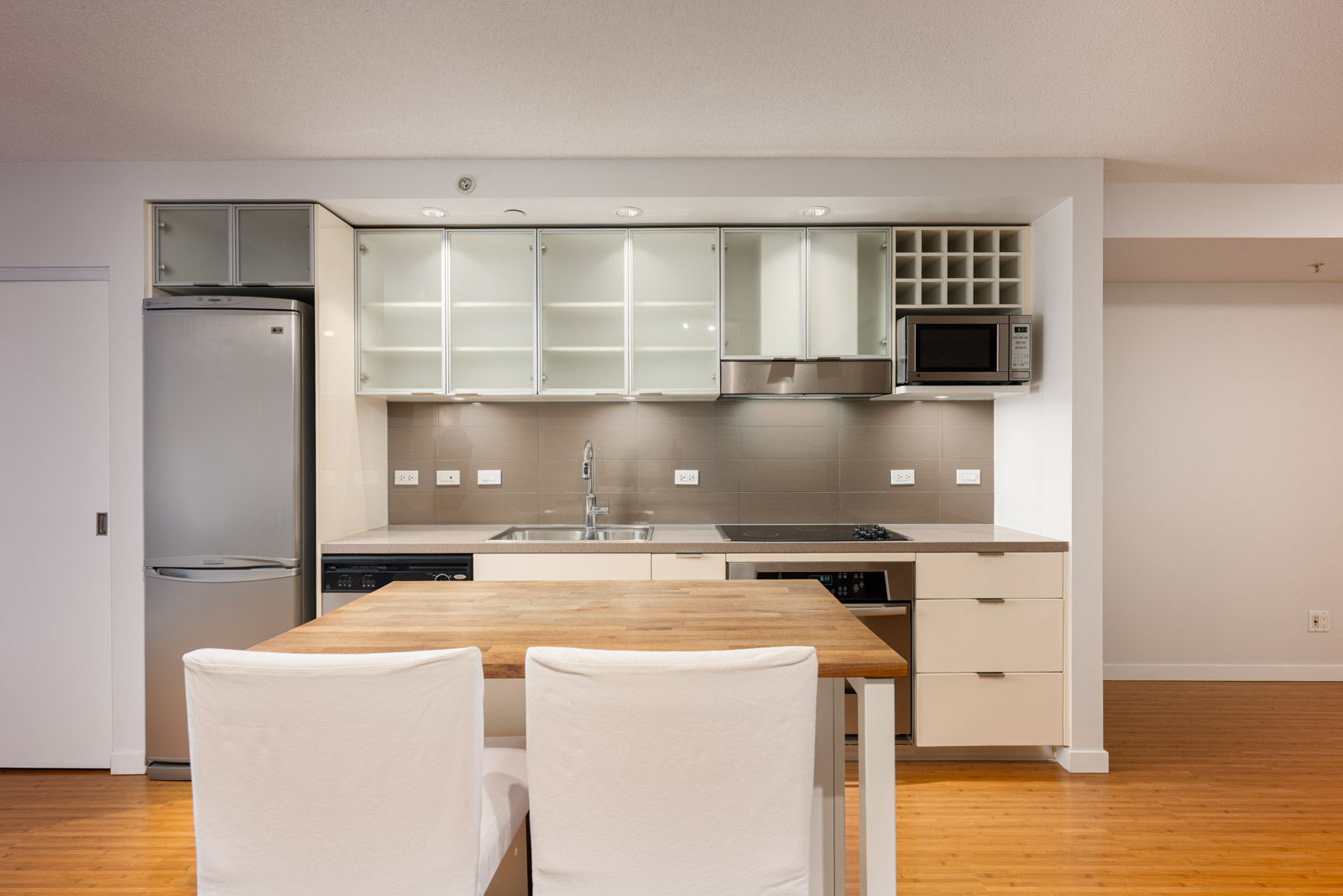 Modern kitchen with white cabinets, stainless steel fridge, microwave, open shelving, and a wooden dining table with two white chairs.