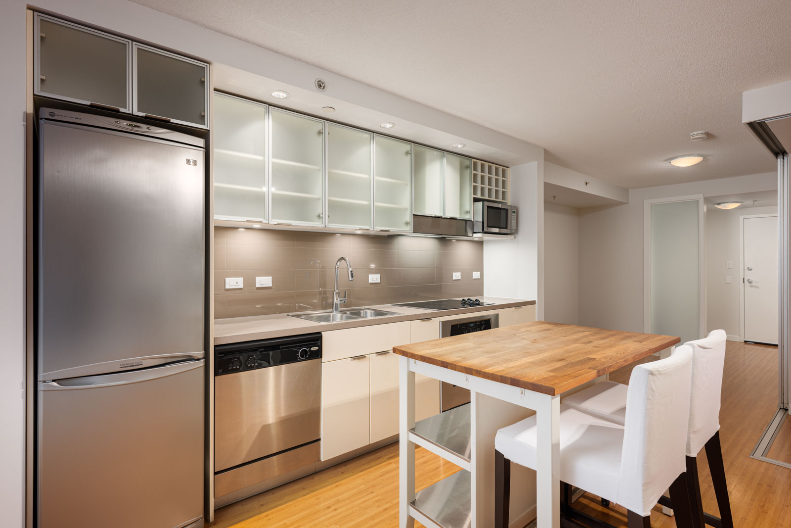 Modern kitchen with stainless steel appliances, glass-front cabinets, a wooden island with two white chairs, and light wood flooring.