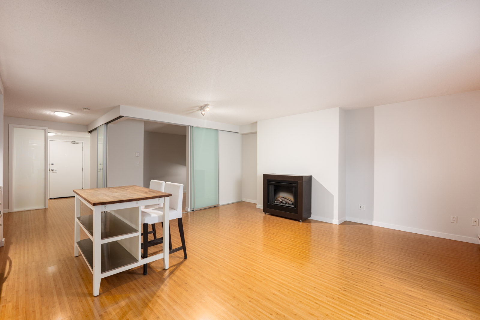 A bright, unfurnished living space with wooden flooring, a small table with two chairs, a modern fireplace, and a white entrance door in the background.