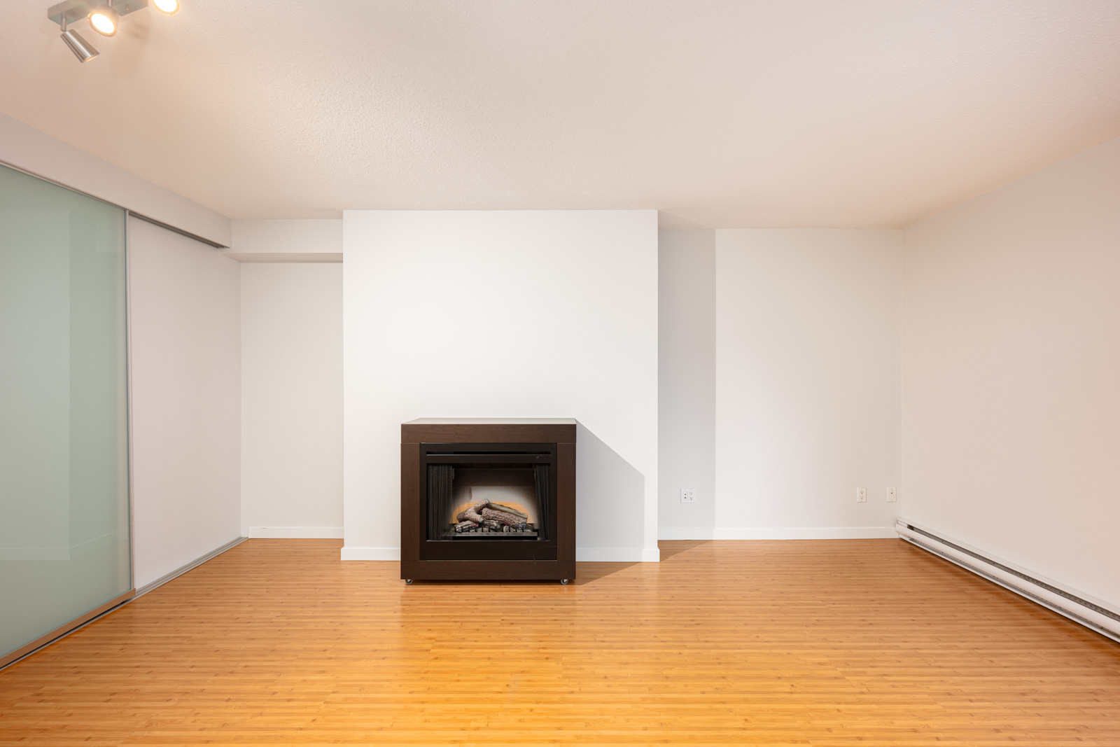 A modern, empty room with light wood flooring, white walls, a dark electric fireplace, and a sliding glass door on the left.