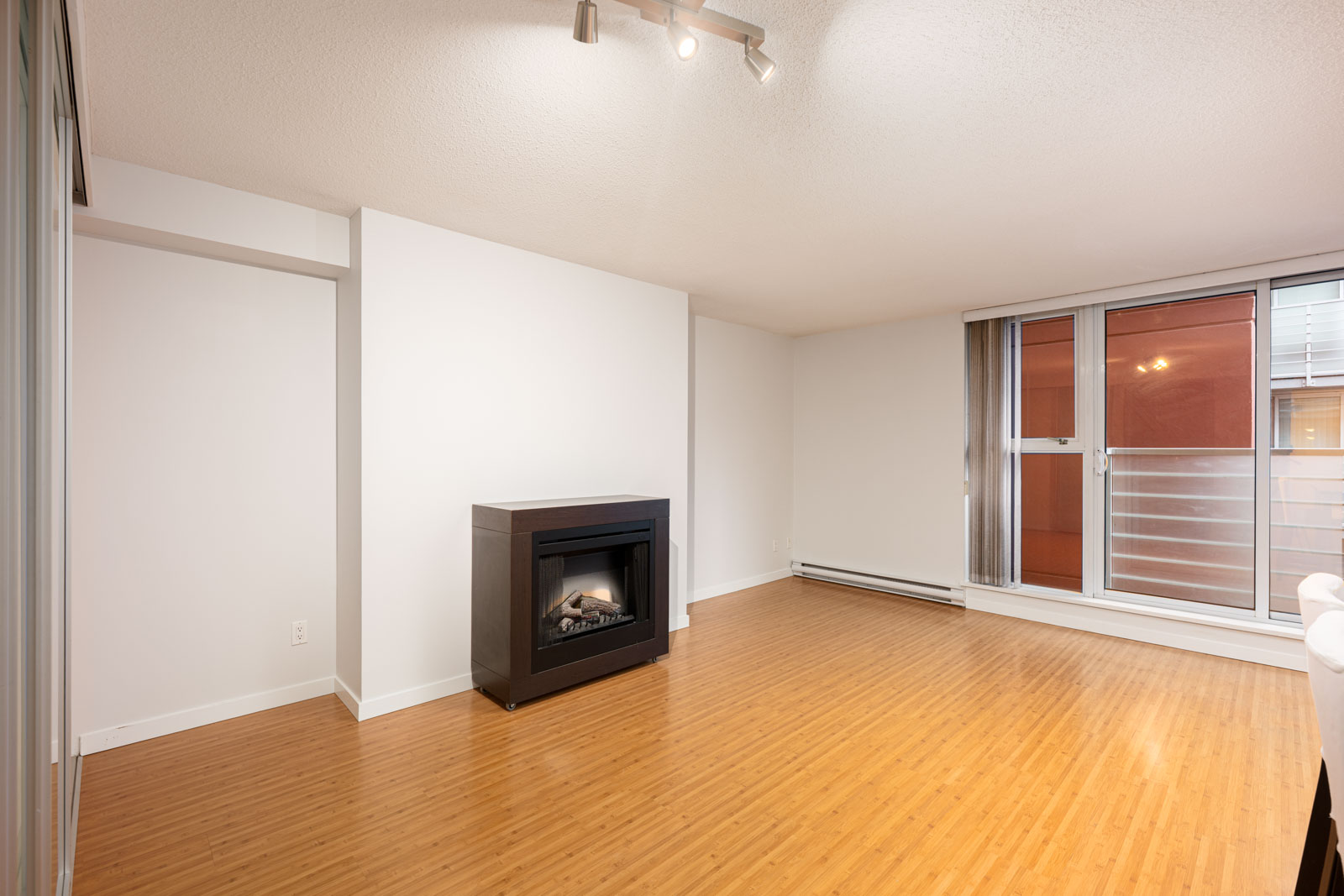 Unfurnished living room with light wood flooring, a modern black fireplace, white walls, ceiling track lights, and large windows with a sliding glass door to a balcony.