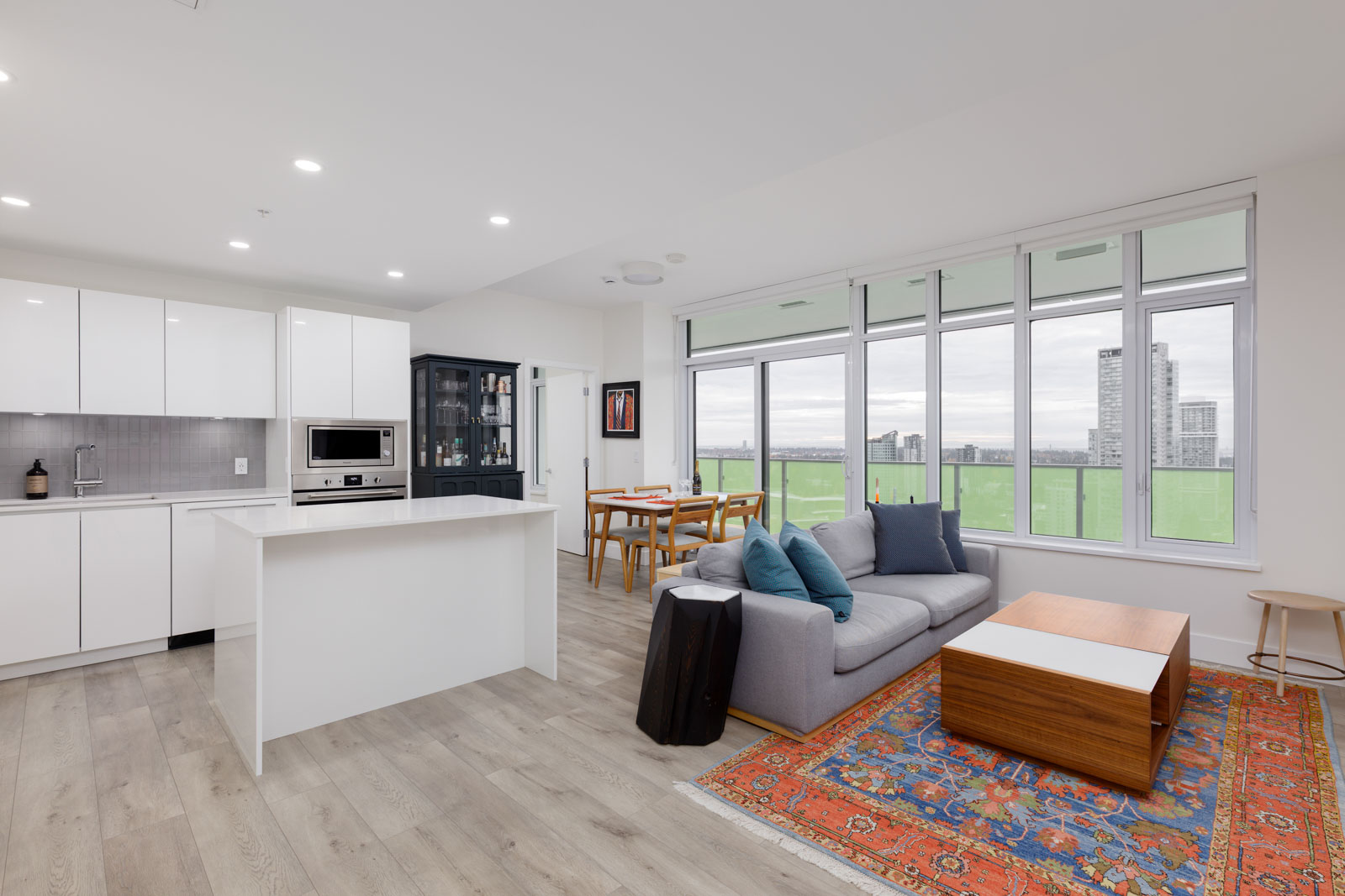 Modern open-plan apartment with white kitchen, small dining area, and living space featuring a gray sofa, wooden coffee table, and large windows overlooking city buildings.