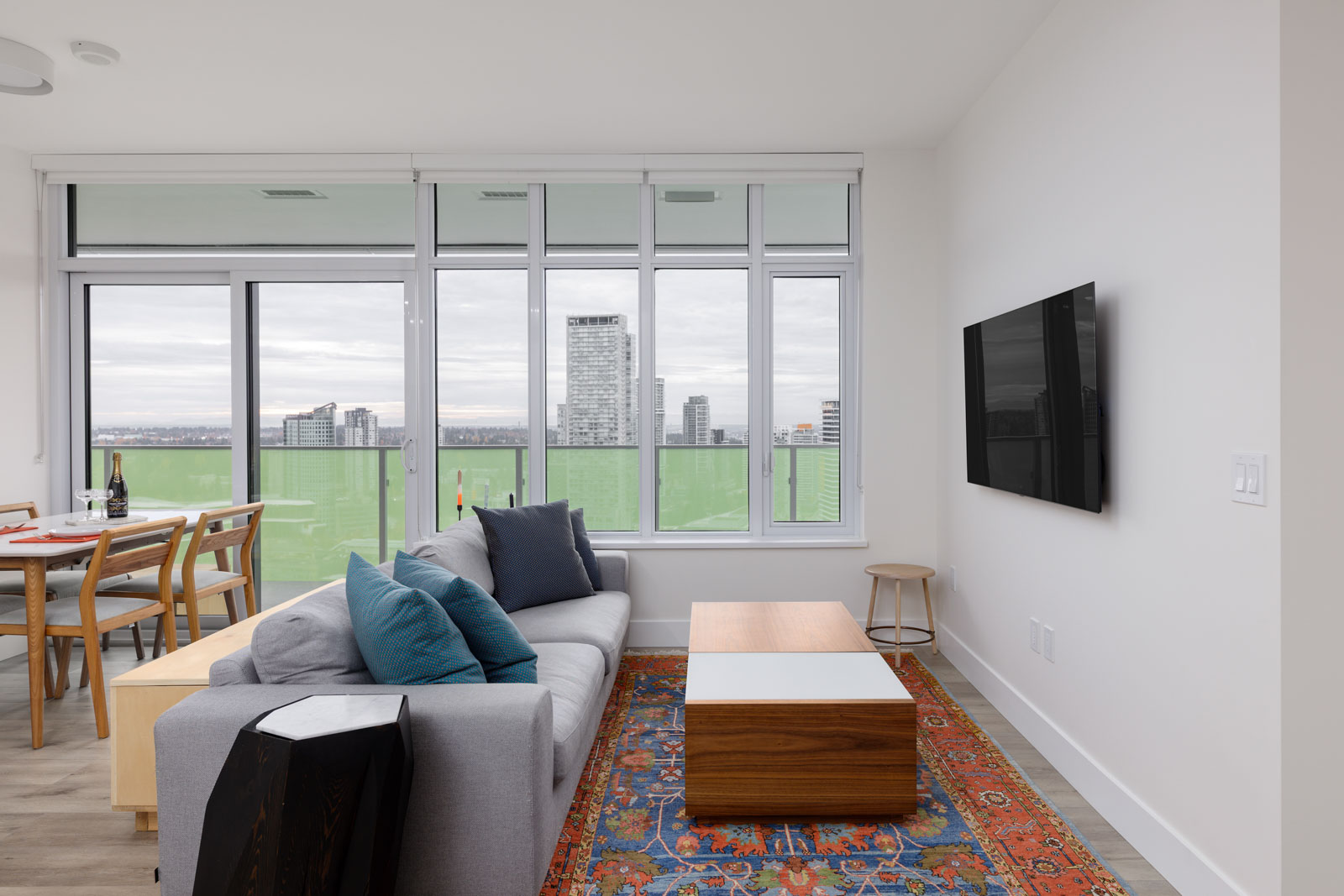 A modern living room with a gray sofa, colorful rug, wall-mounted TV, wooden coffee table, dining area, and large windows overlooking city buildings.