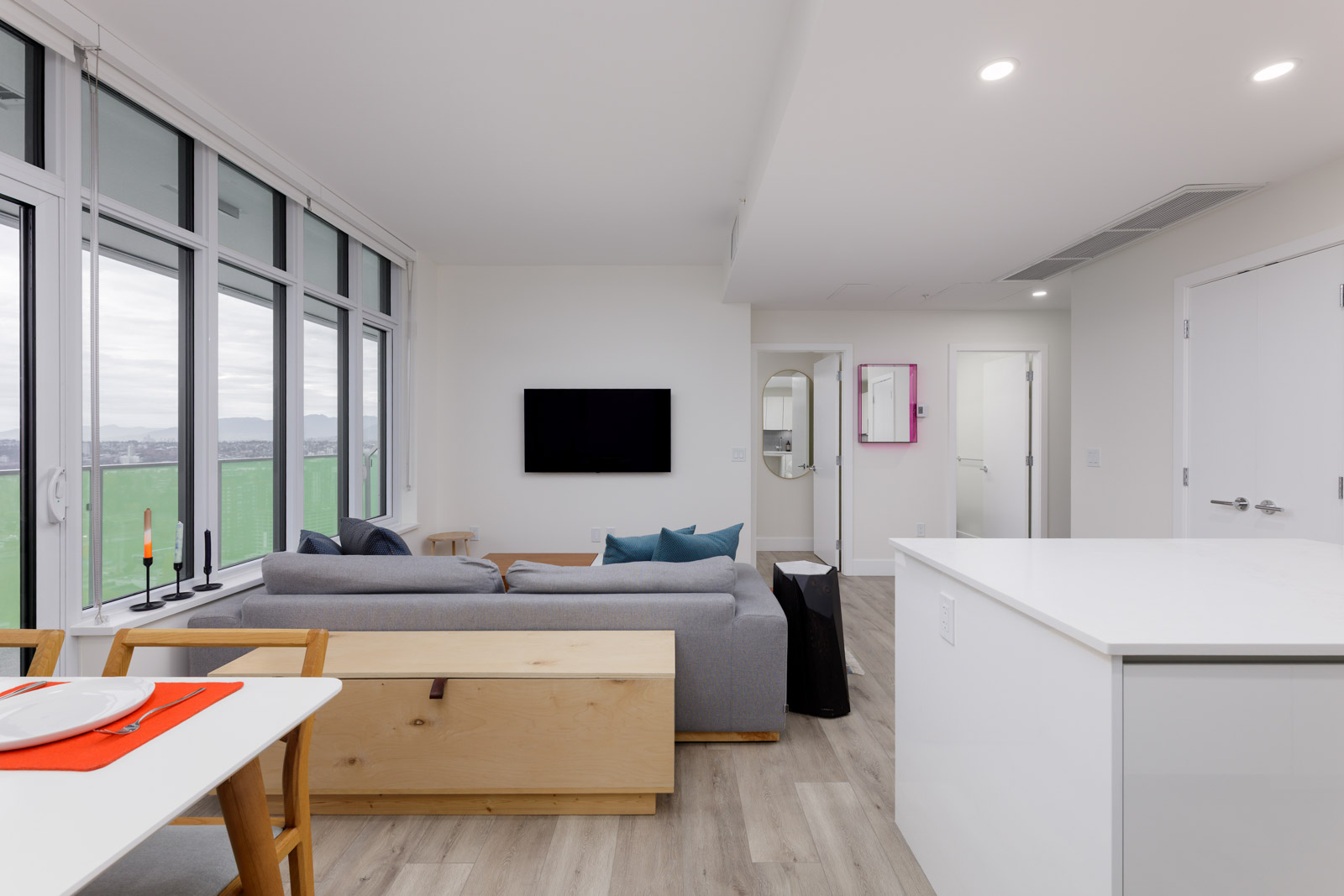 Modern living room and kitchen area with a gray sofa, wall-mounted TV, large windows, and light wood flooring. Dining table and kitchen counter are in the foreground.