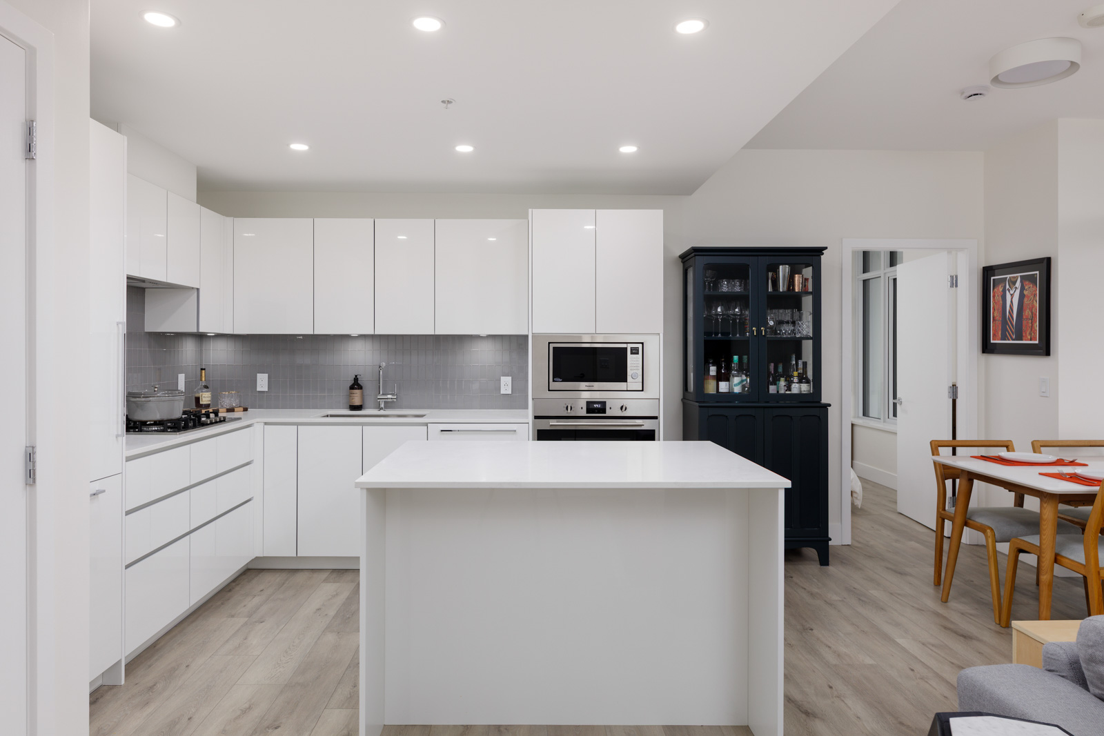 Modern kitchen with white cabinets and island, gray backsplash, built-in appliances, and adjacent dining area with a black cabinet and framed art on the wall.