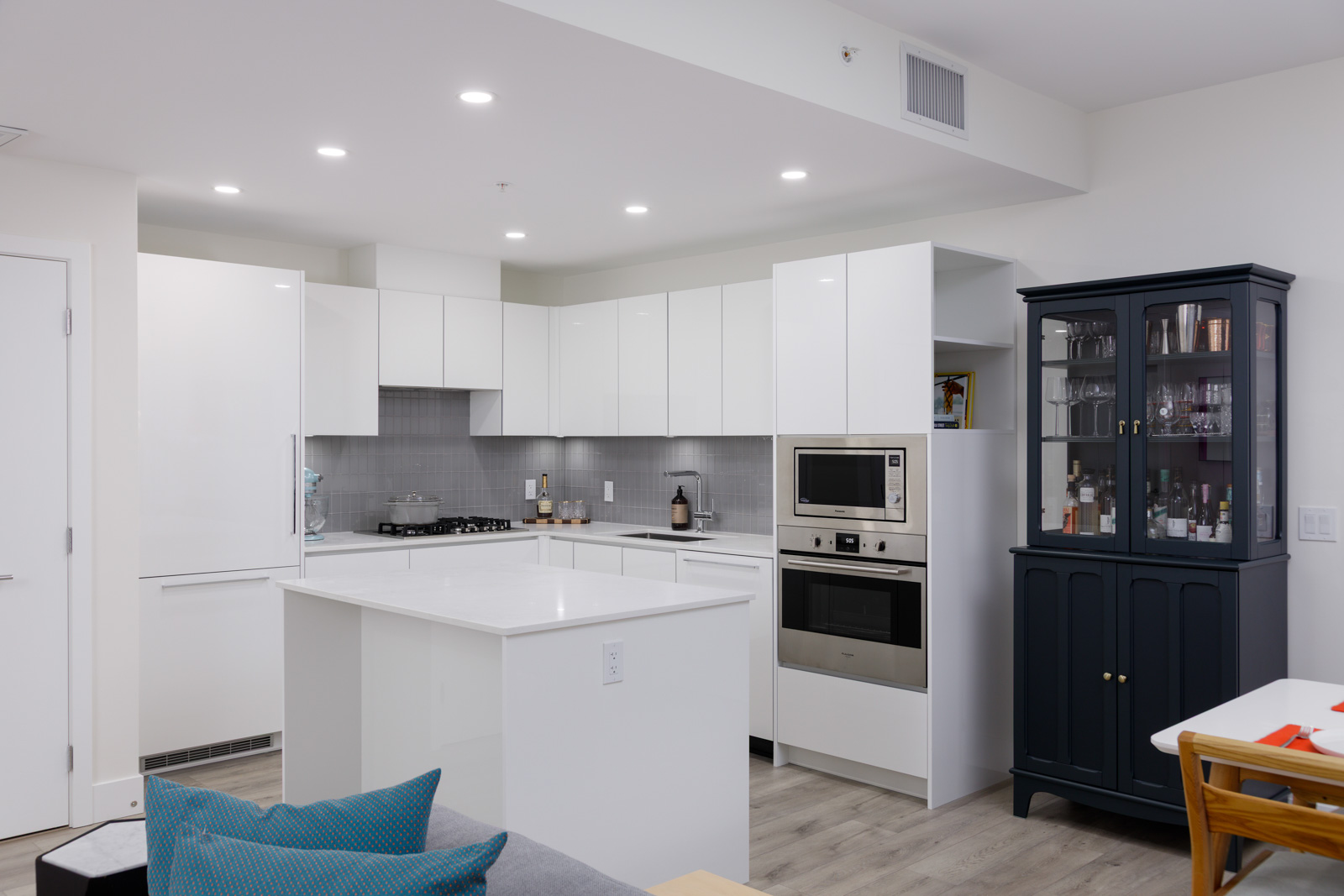 Modern kitchen with white cabinets, built-in oven, microwave, and a black glass-fronted cabinet. Gray backsplash, white countertops, and wooden flooring are visible.