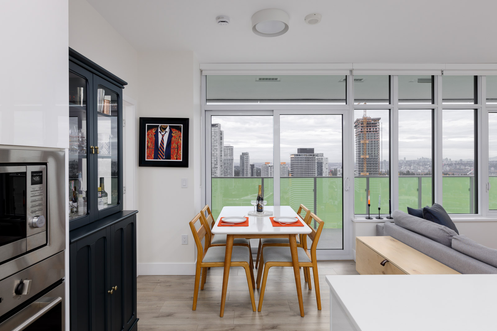 A modern dining area with a white table set for two, wooden chairs, glass doors leading to a balcony, and a cityscape view in the background.