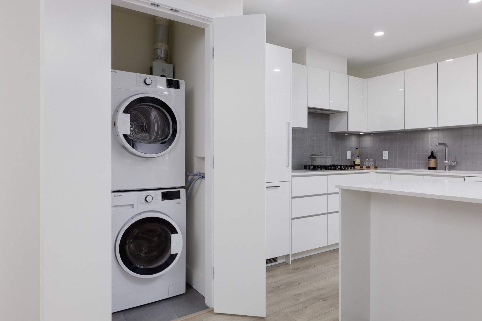A modern laundry area with a stacked washer and dryer is located in a closet next to a contemporary white kitchen.
