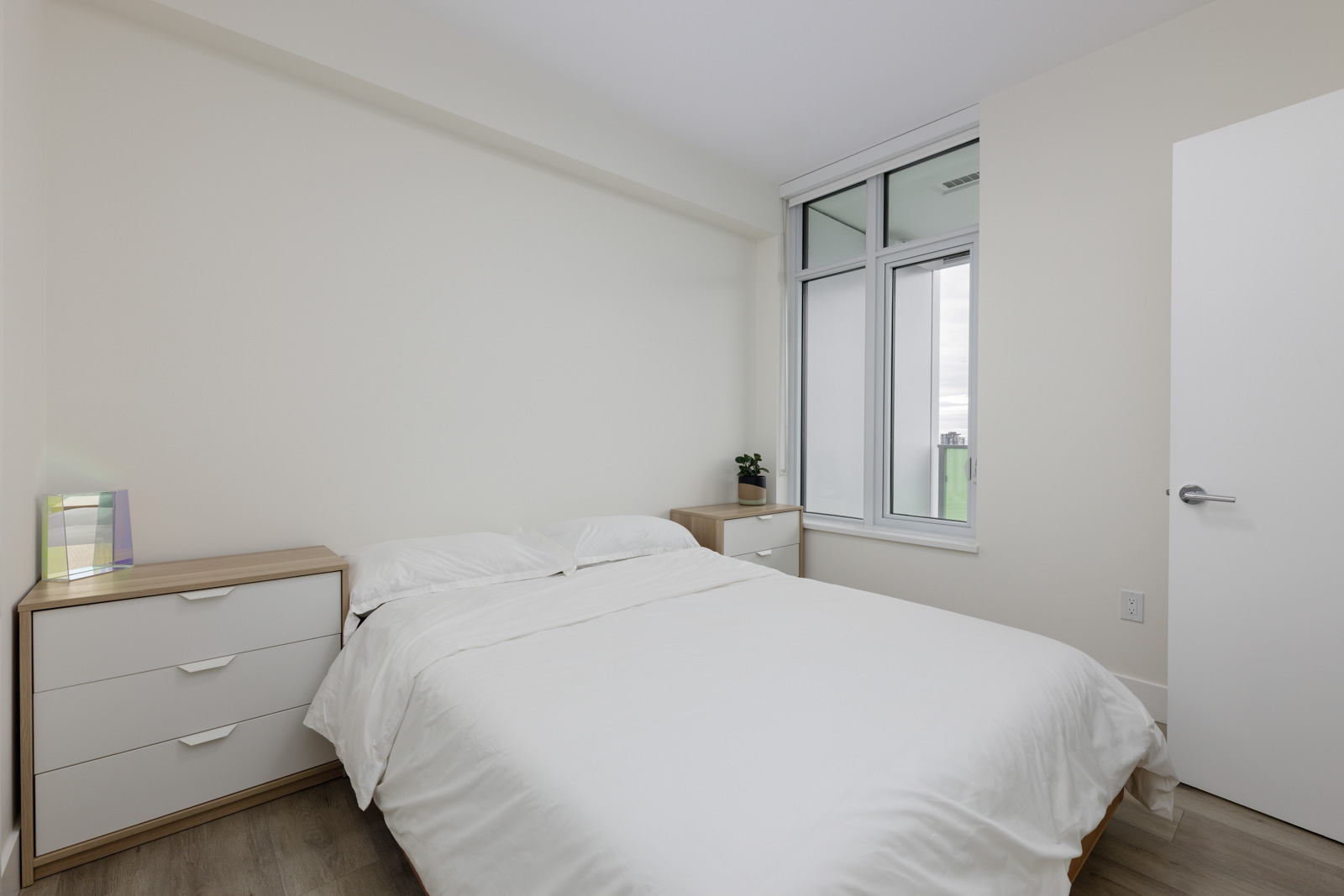 A small, minimalist bedroom with a neatly made bed, two wooden nightstands, a closed white door, and a window letting in natural light.