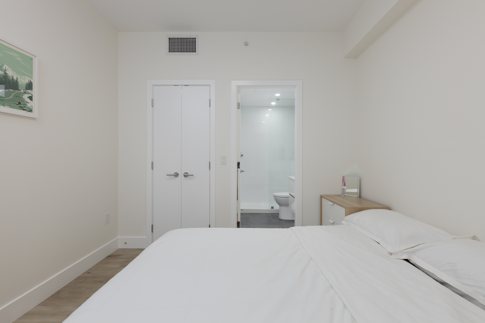 Minimalist bedroom with a neatly made bed, a small bedside table, white walls, double closet doors, and an open door to a bathroom.