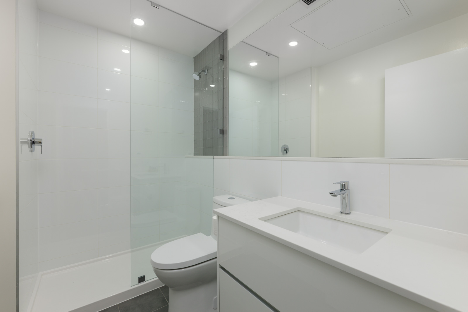 Modern bathroom with a glass-enclosed shower, white sink vanity, toilet, large mirror, and white tiled walls.