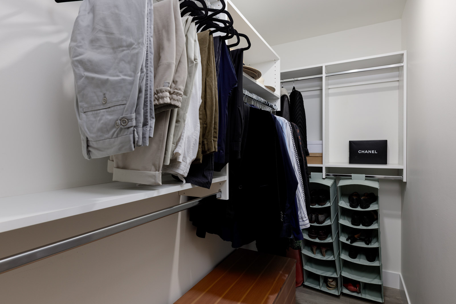 A modern walk-in closet with hanging clothes, a shoe rack filled with shoes, shelves, and a Chanel box on a shelf in the back.