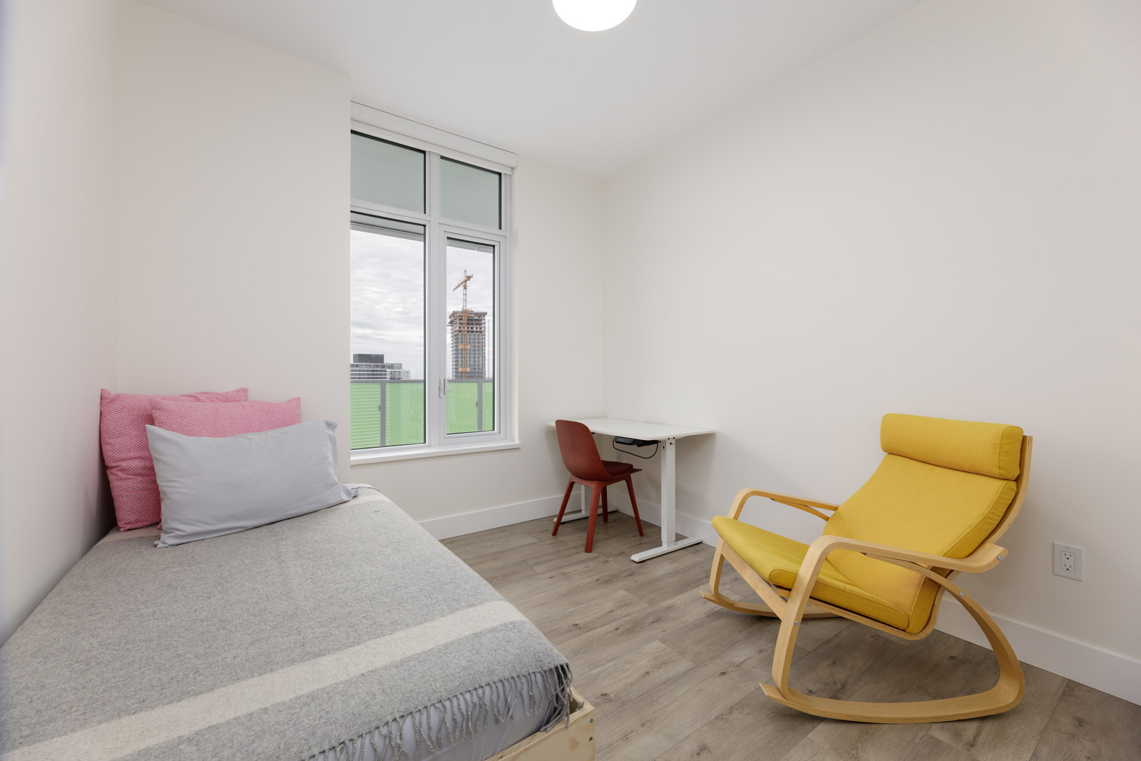 Minimalist bedroom with a single bed, pink and gray bedding, a small desk with a red chair, a yellow armchair, and a window showing a cityscape under construction.