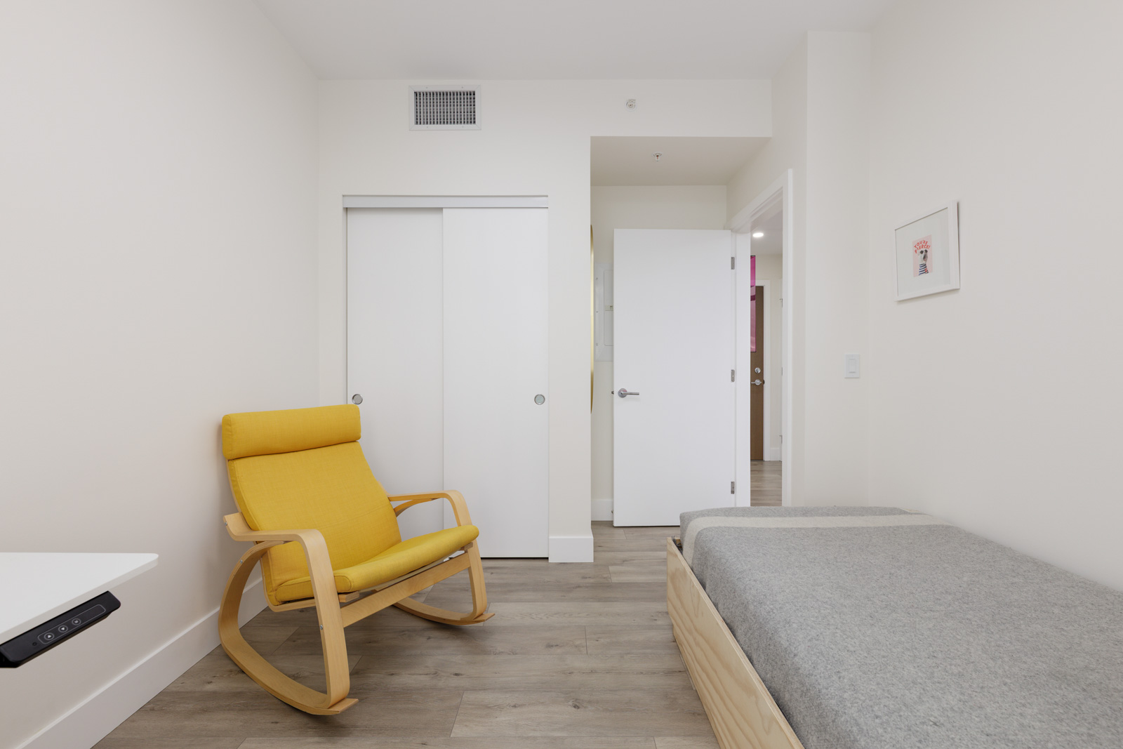 Minimalist bedroom with a wooden bed, gray blanket, yellow rocking chair, white wall, closed closet doors, and an open door leading to a hallway.
