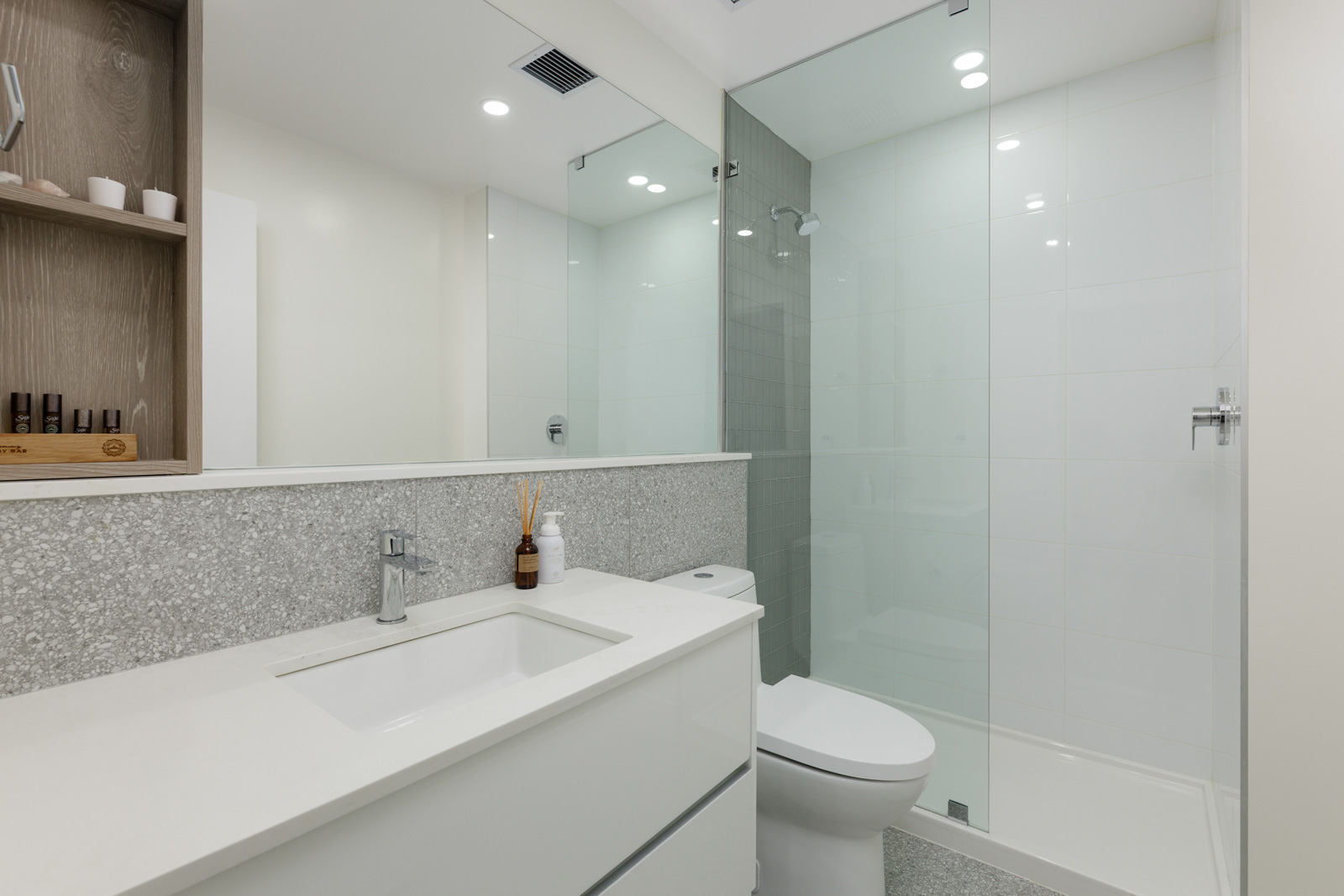 Modern bathroom with a white vanity, sink, toilet, glass shower enclosure, large mirror, and recessed shelf with toiletries.