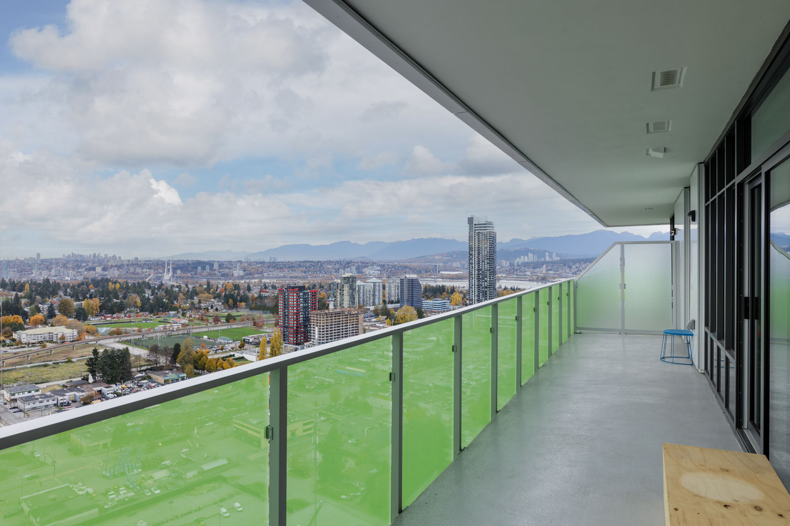 A high-rise balcony with green-tinted glass railing overlooks a cityscape, distant mountains, and cloudy sky.