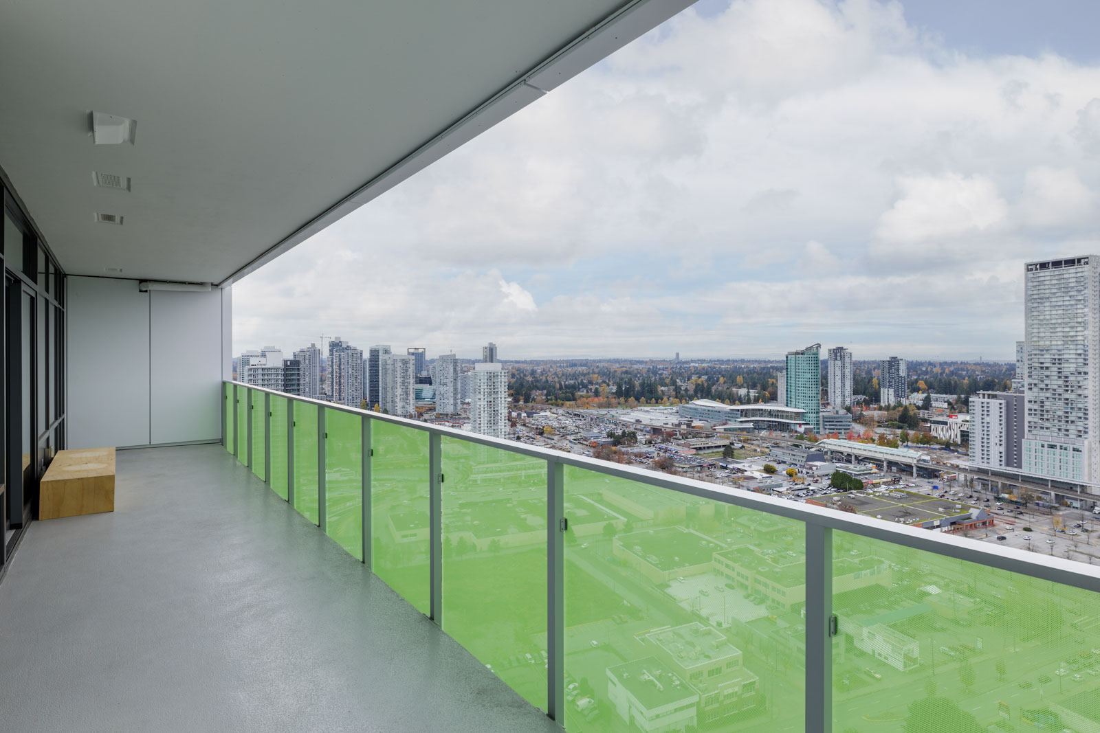 A high-rise balcony with a green-tinted glass railing overlooks a cityscape with tall buildings and a cloudy sky.
