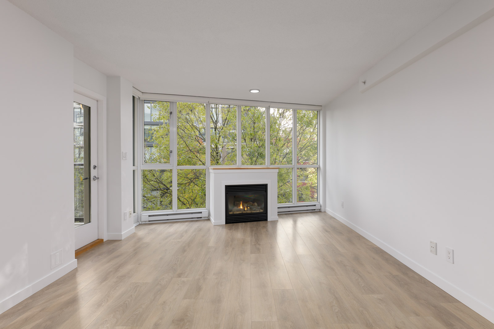 Empty living room with light wood flooring, large windows with a view of green trees, a white wall, and a fireplace in the center.