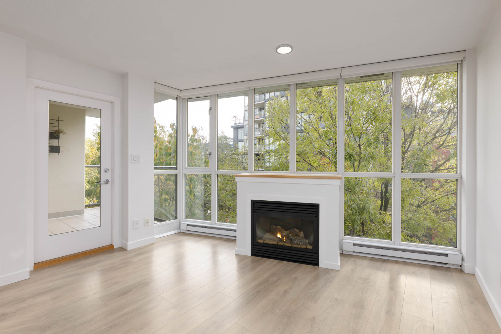 Bright, empty room with light wood flooring, a glass door, large windows, and a fireplace beneath a shelf. Trees and a building are visible outside.