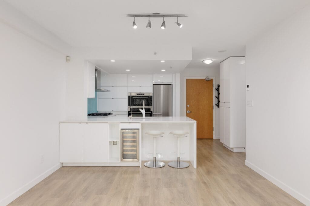 Modern white kitchen with an island, two bar stools, built-in wine cooler, stainless steel appliances, and light wood flooring.