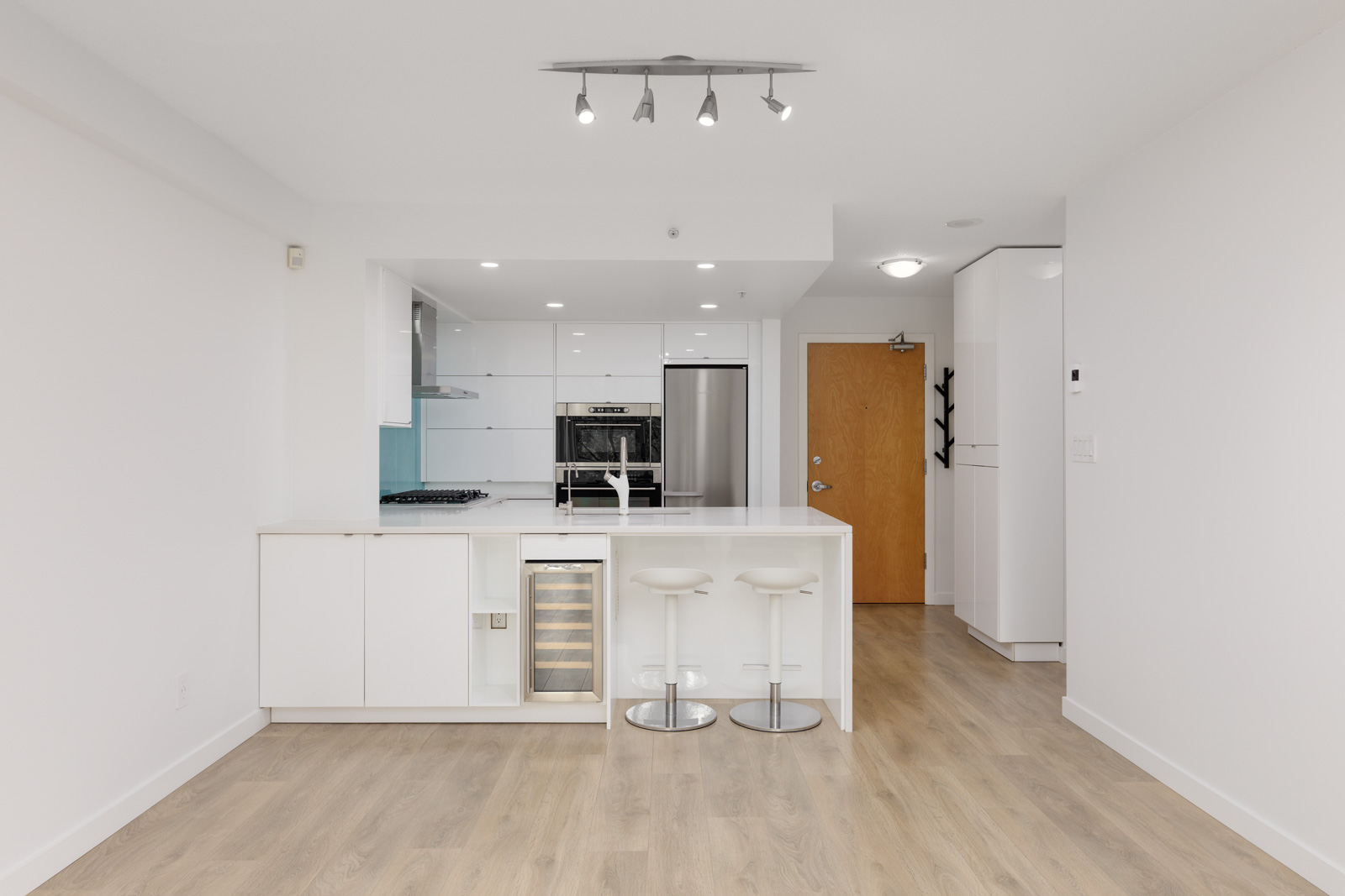 Modern white kitchen with an island, two bar stools, built-in wine cooler, stainless steel appliances, and light wood flooring.