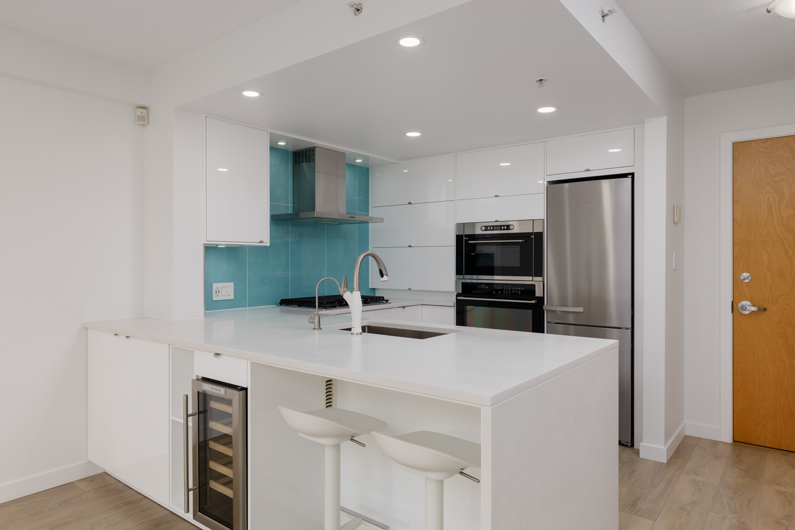 Modern kitchen with white cabinetry, stainless steel appliances, a glass cooktop, turquoise backsplash, and a white island with two bar stools.