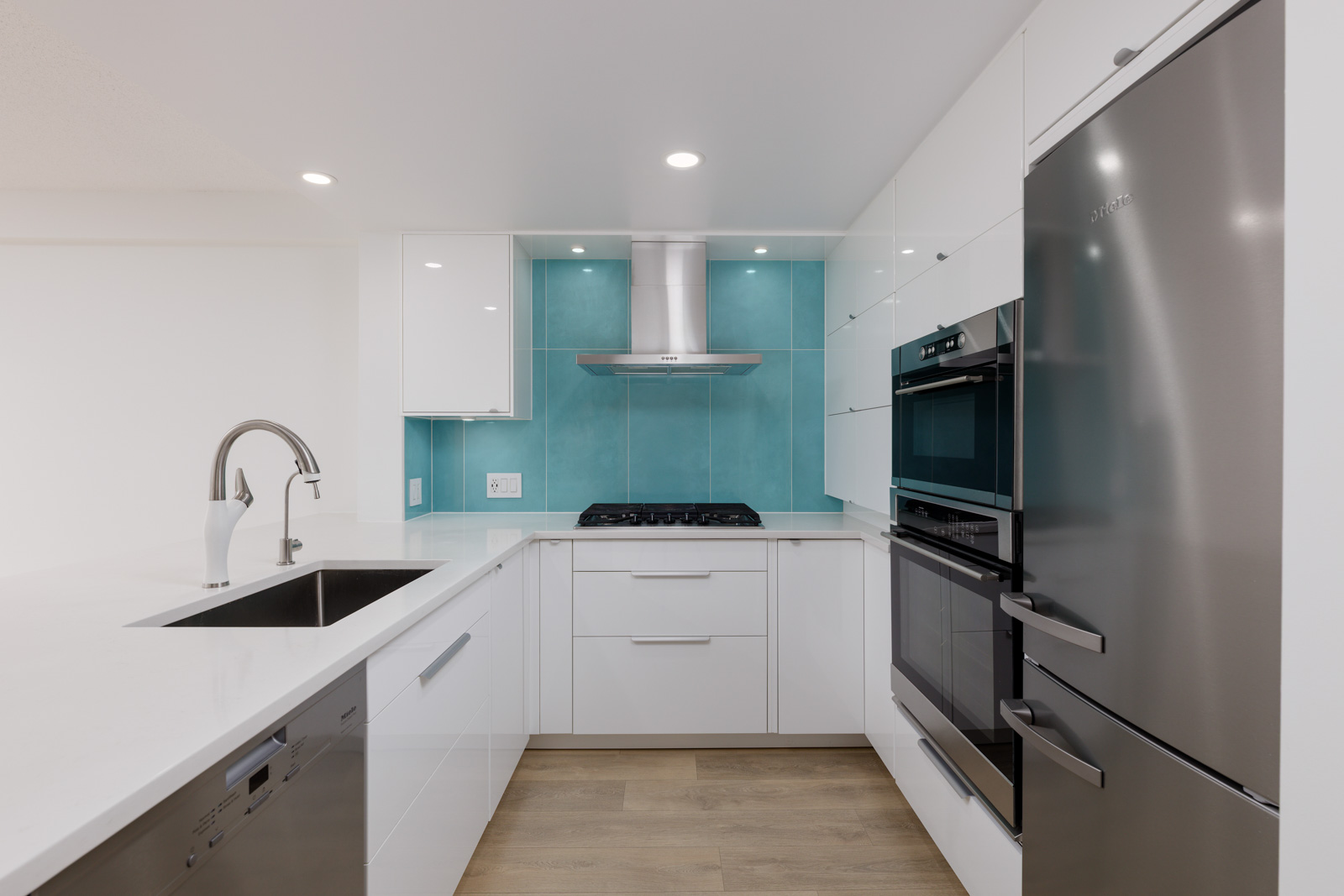 Modern kitchen with white cabinets, stainless steel appliances, a built-in oven, gas cooktop, and turquoise backsplash under a range hood.