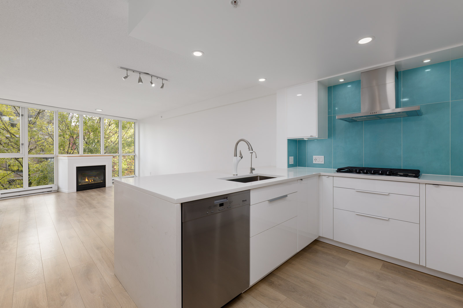 Modern kitchen with white cabinets, stainless steel appliances, turquoise backsplash, and an island; open layout leads to living area with large windows and a fireplace.