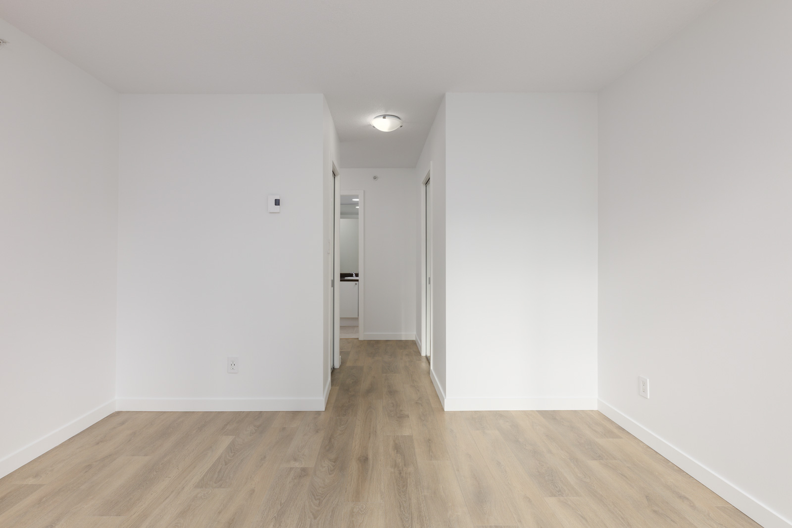 Empty room with white walls, light wood flooring, and a hallway leading to doorways. Ceiling light and thermostat are visible.