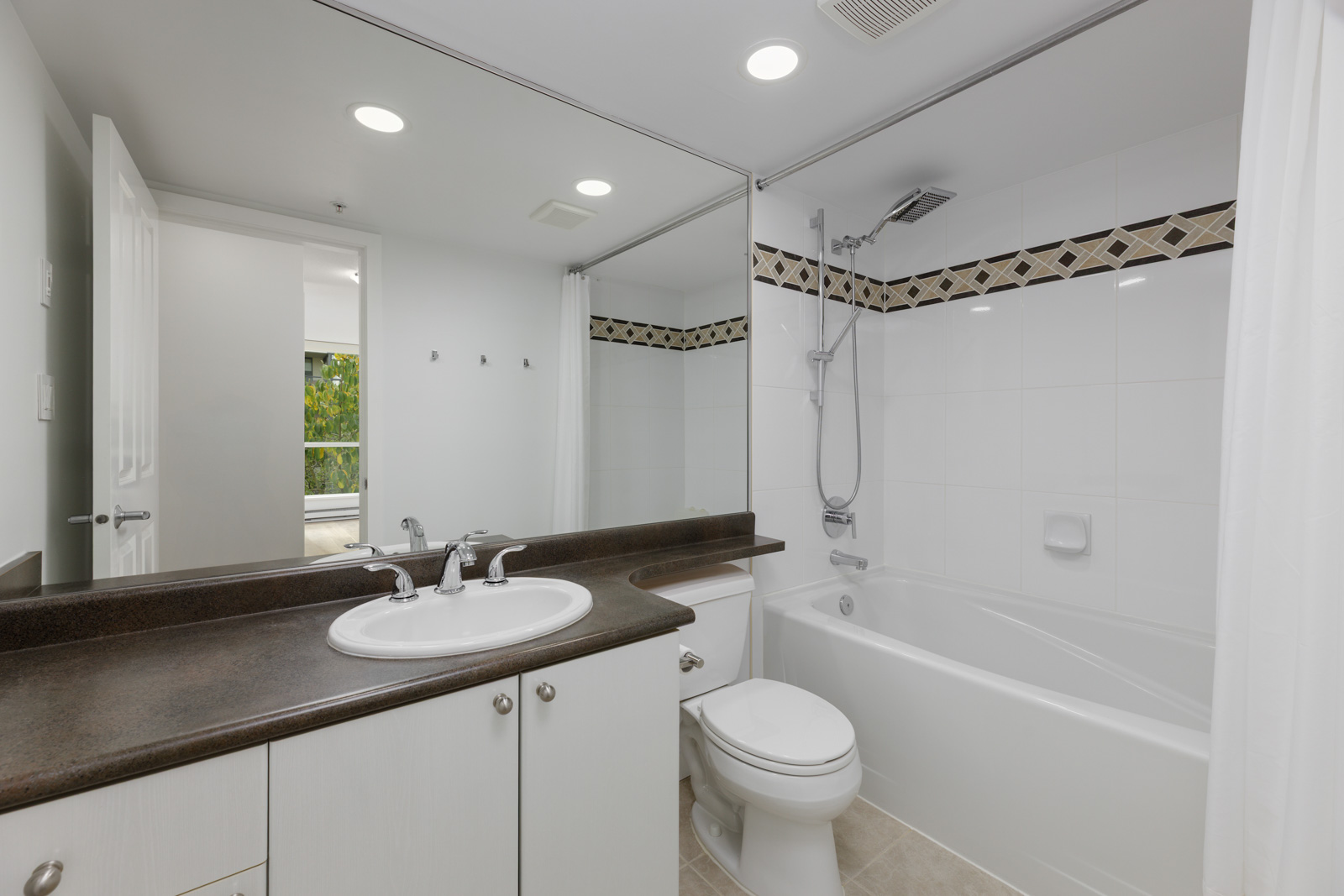 Modern bathroom with a large mirror, white sink and cabinet, dark countertop, toilet, and a bathtub with a shower and decorative tile border.