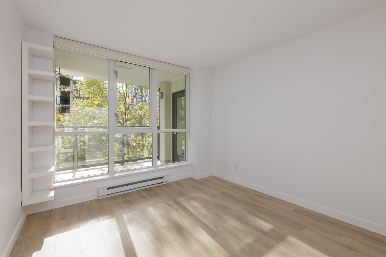 A bright empty room with light wood flooring, white walls, built-in shelving, and large windows overlooking a balcony and trees outside.