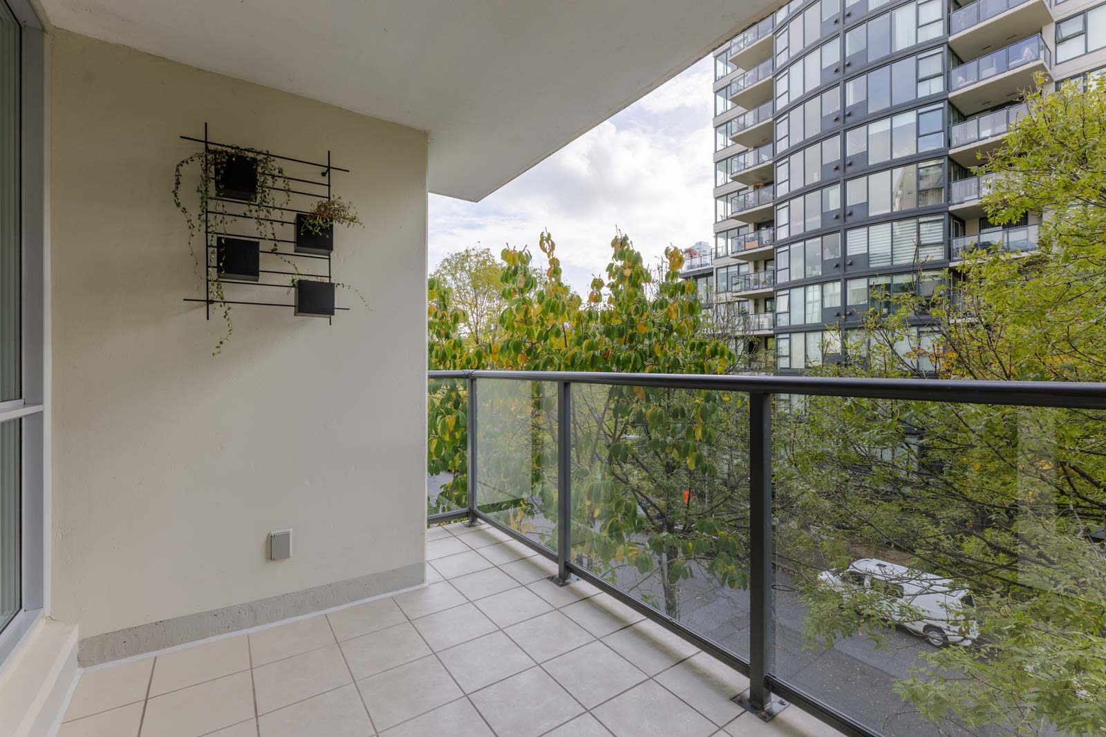 A tiled balcony with glass railing, a wall-mounted planter with a few plants, and a view of trees and a modern apartment building across the street.