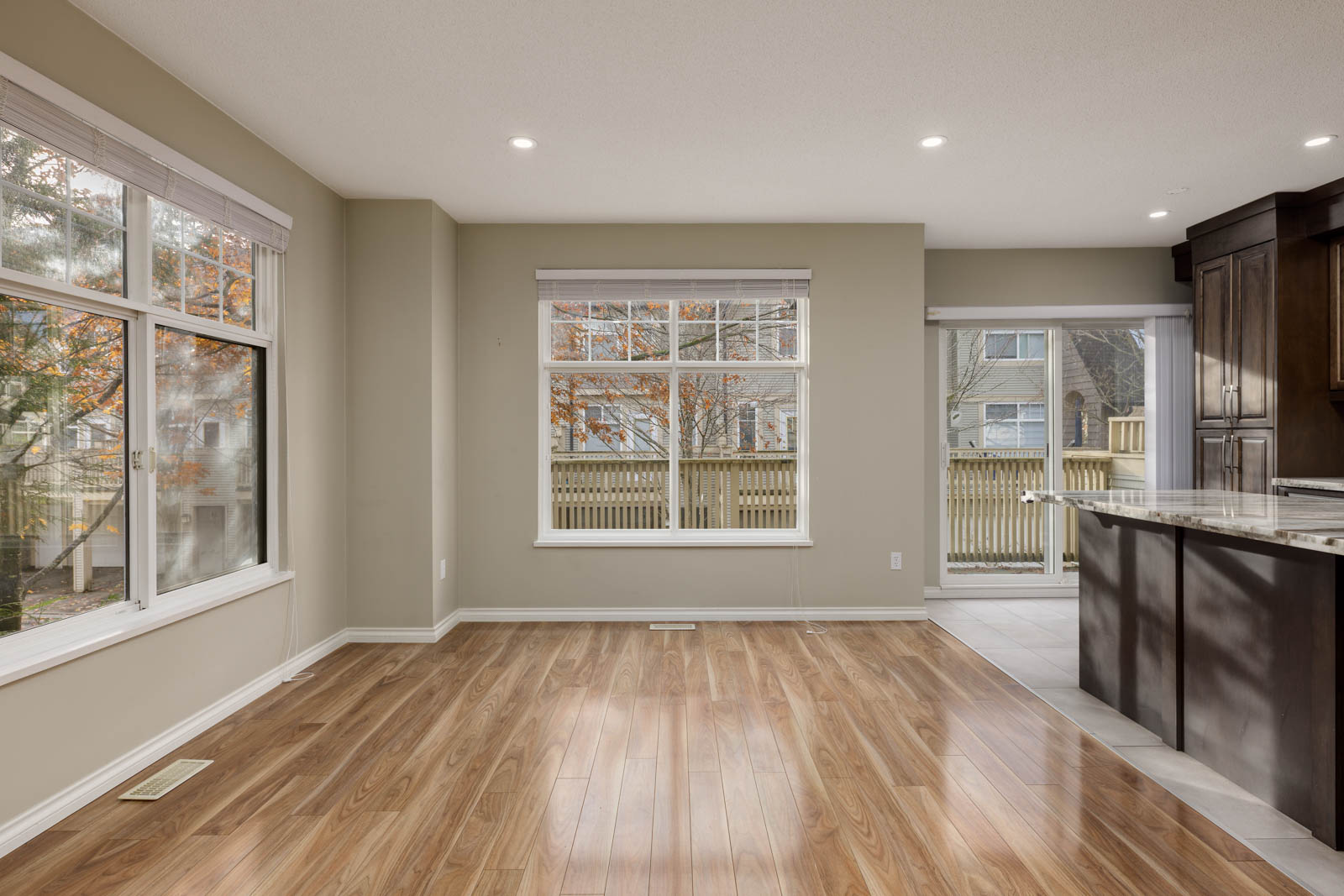 Empty room with light brown wood flooring, large windows with white blinds, beige walls, and partial view of a kitchen with dark cabinets and granite countertop.